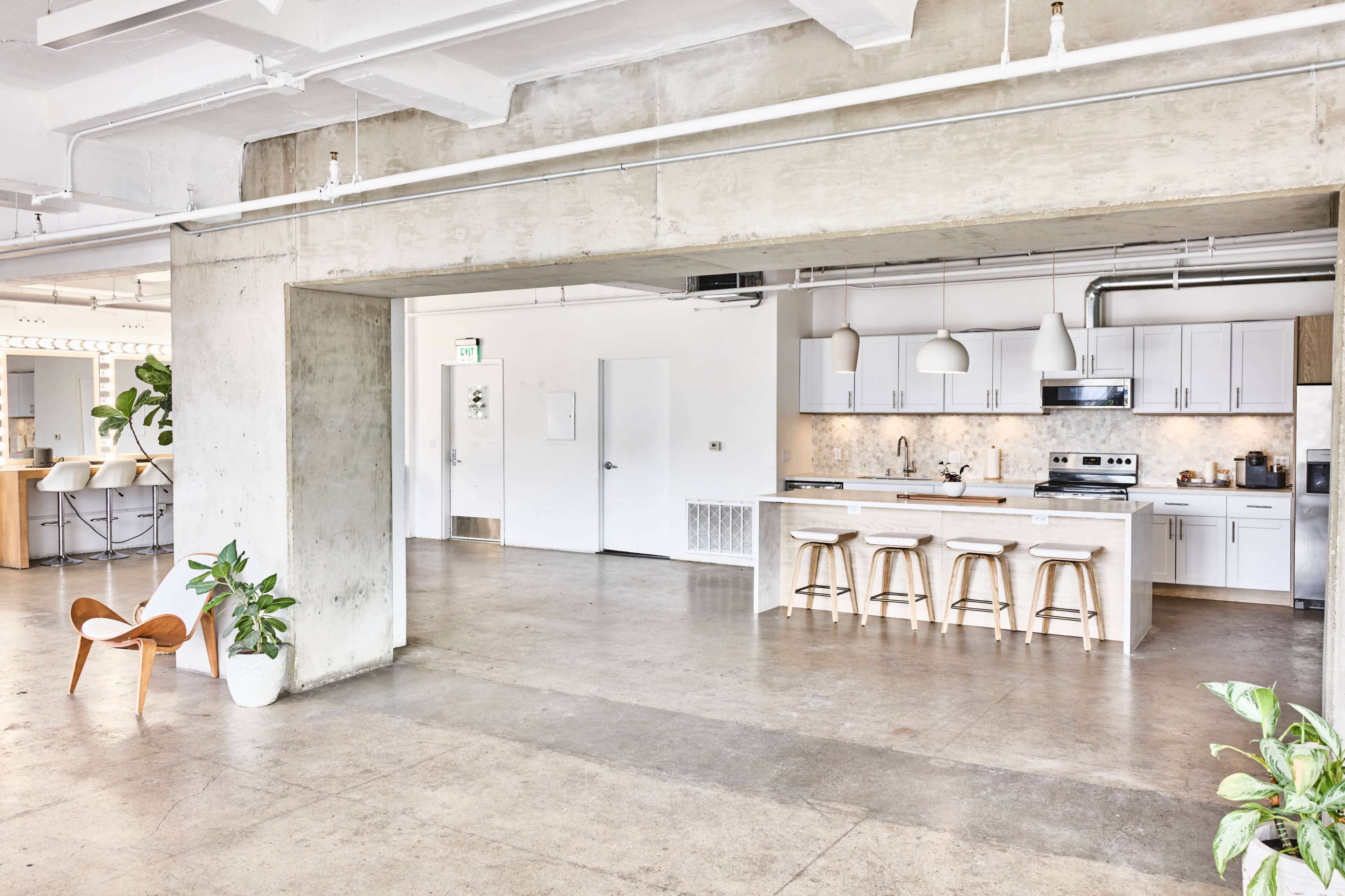 The image shows a modern, open-concept kitchen area with concrete walls and a spacious dining space featuring a wooden table and chairs.