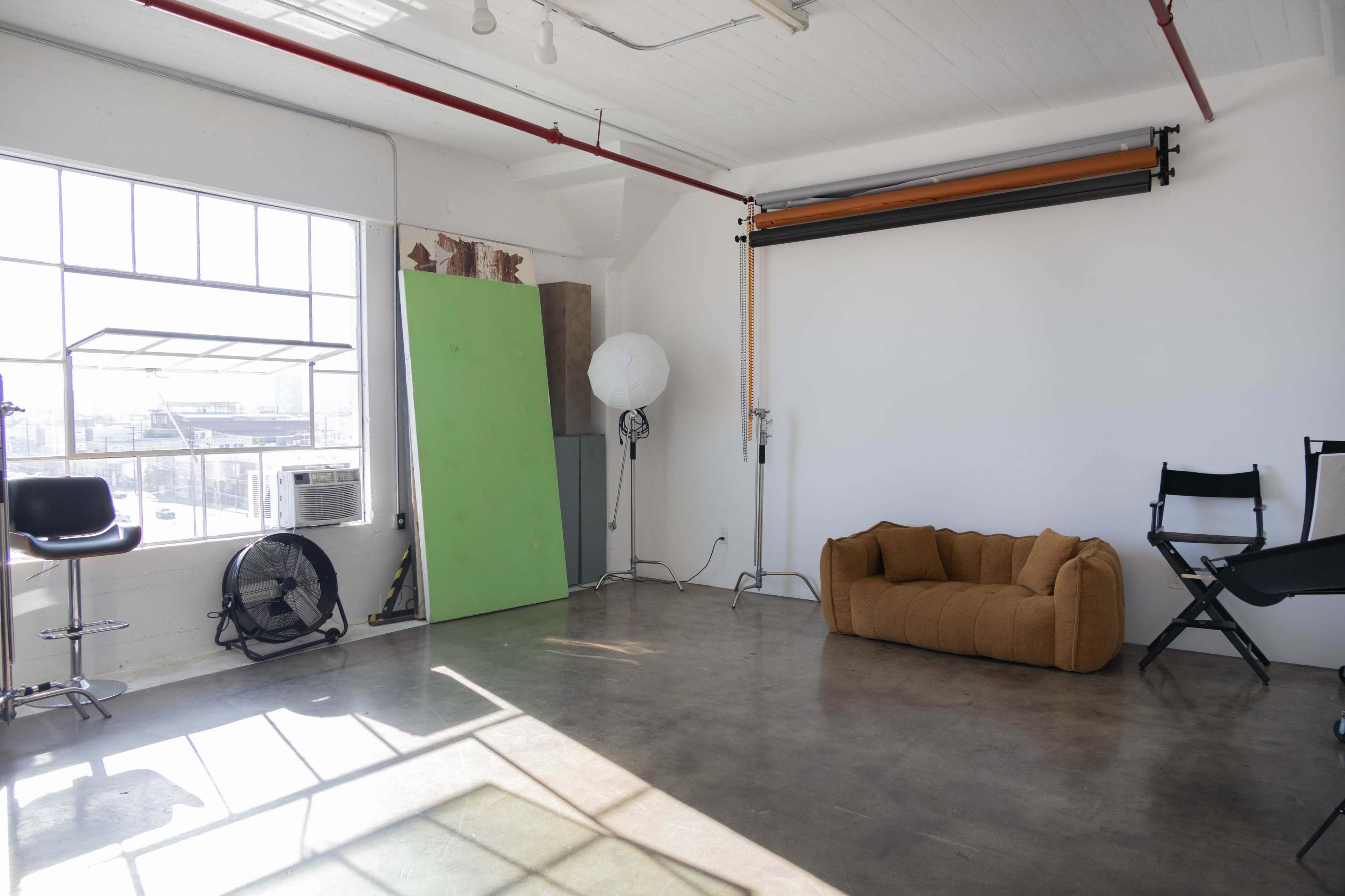 The image shows an empty studio space with large windows, a couch, and various photography equipment set against white walls.