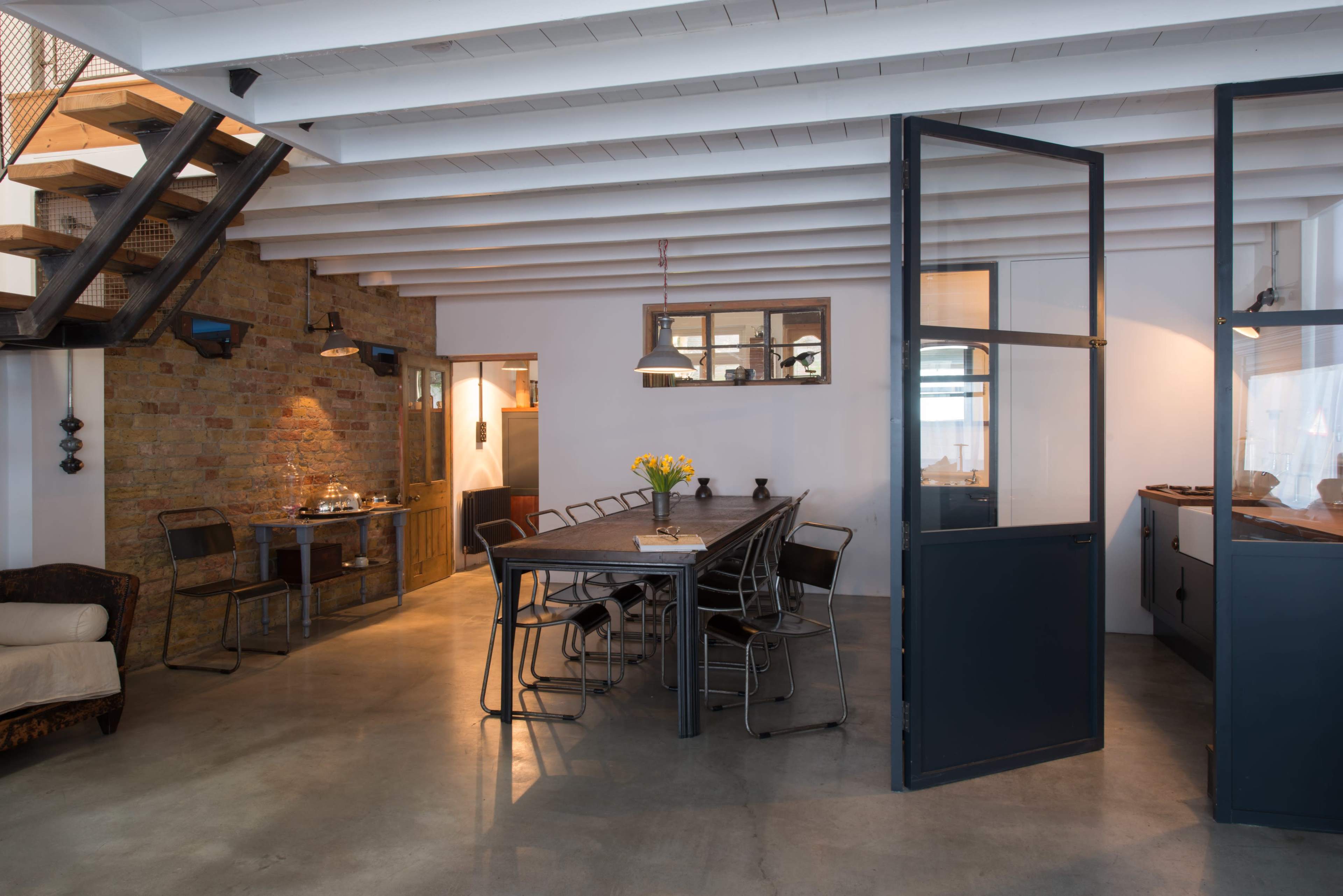 The image shows a modern, open-concept interior space featuring a dining table surrounded by chairs, a staircase, and a mix of brick and white walls.