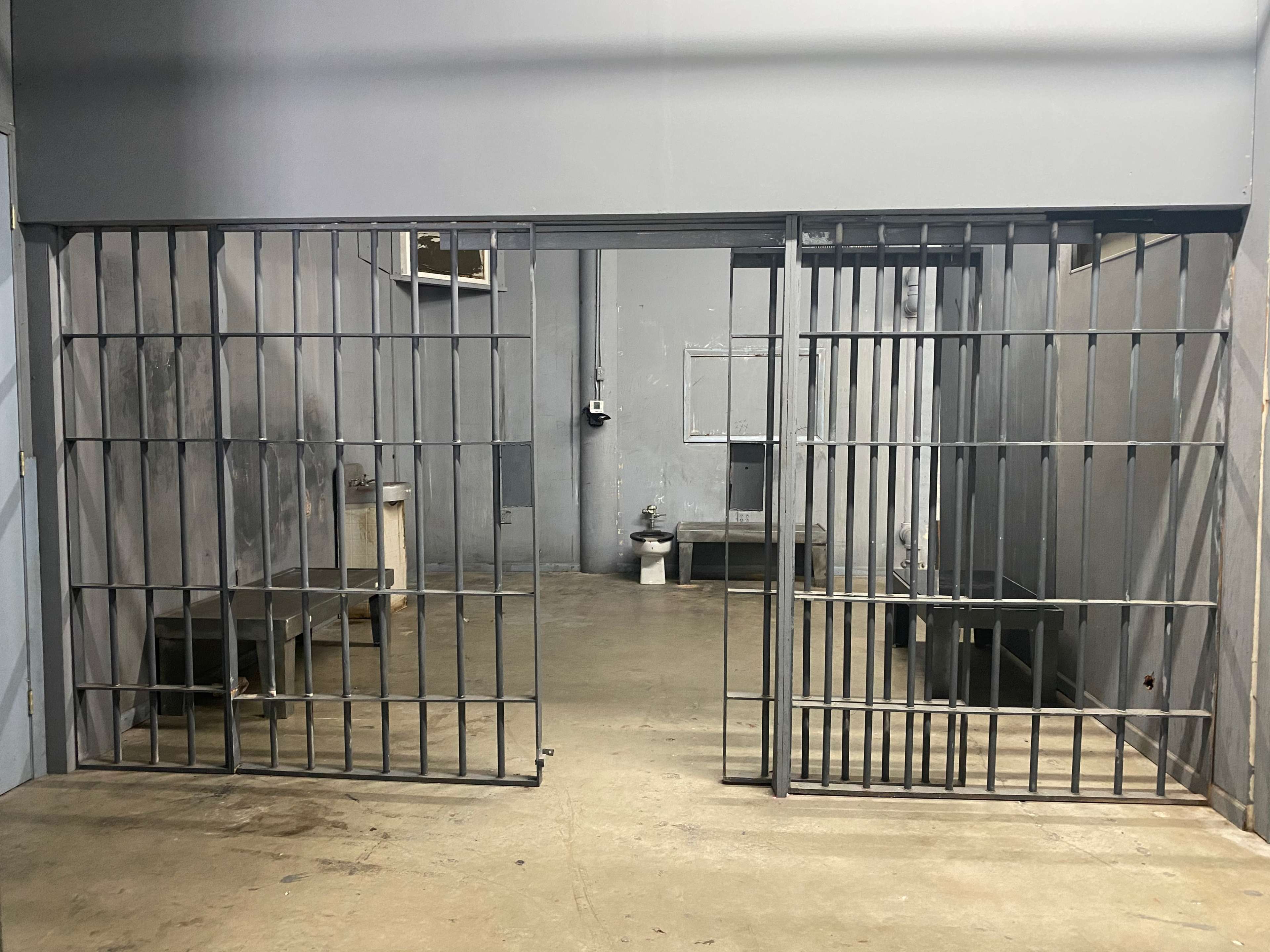 The image shows two open jail cell doors in a dimly lit, empty room with concrete floors and minimal furnishings.