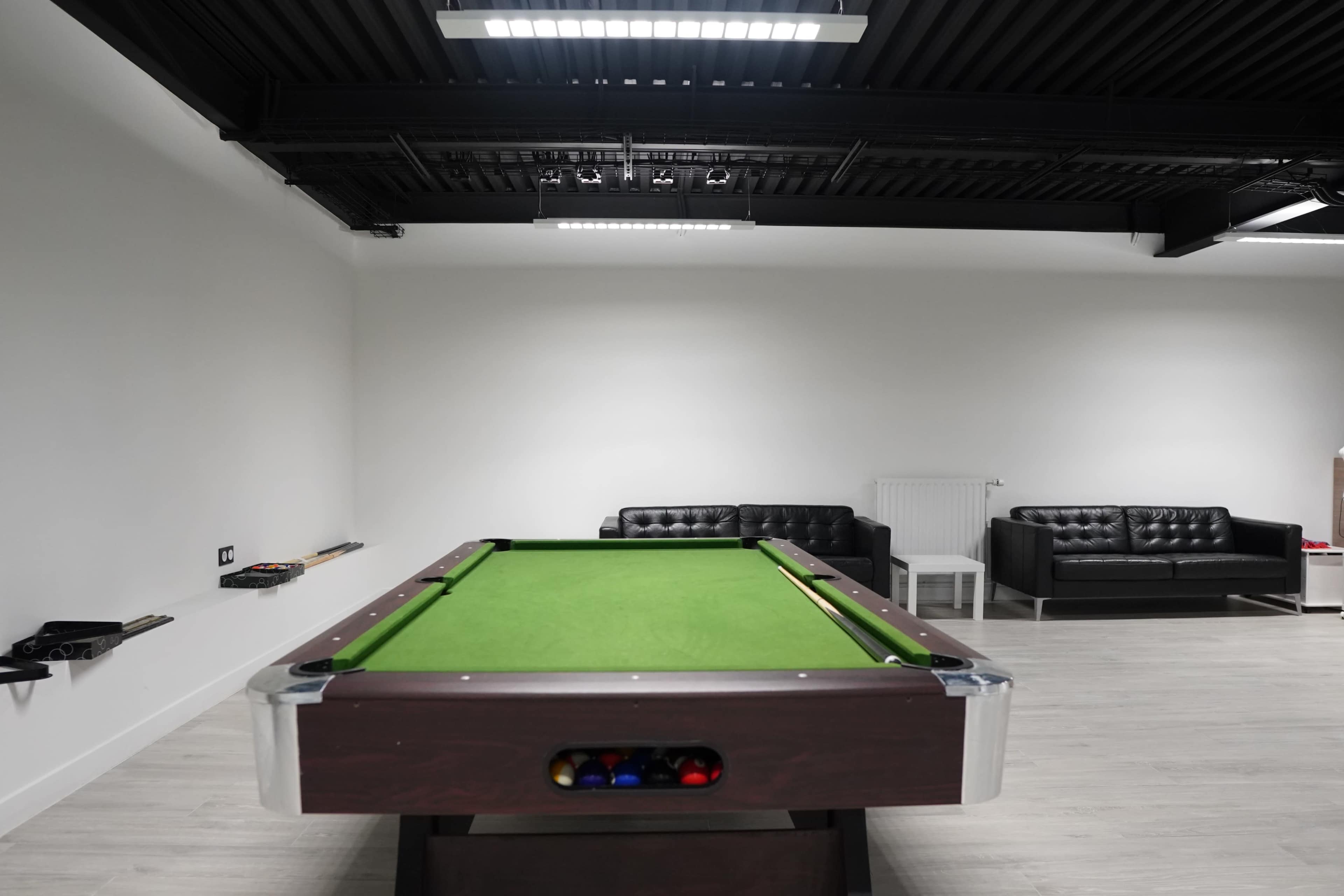 A well-lit room features a green billiards table with a rack of balls, flanked by two black leather sofas.