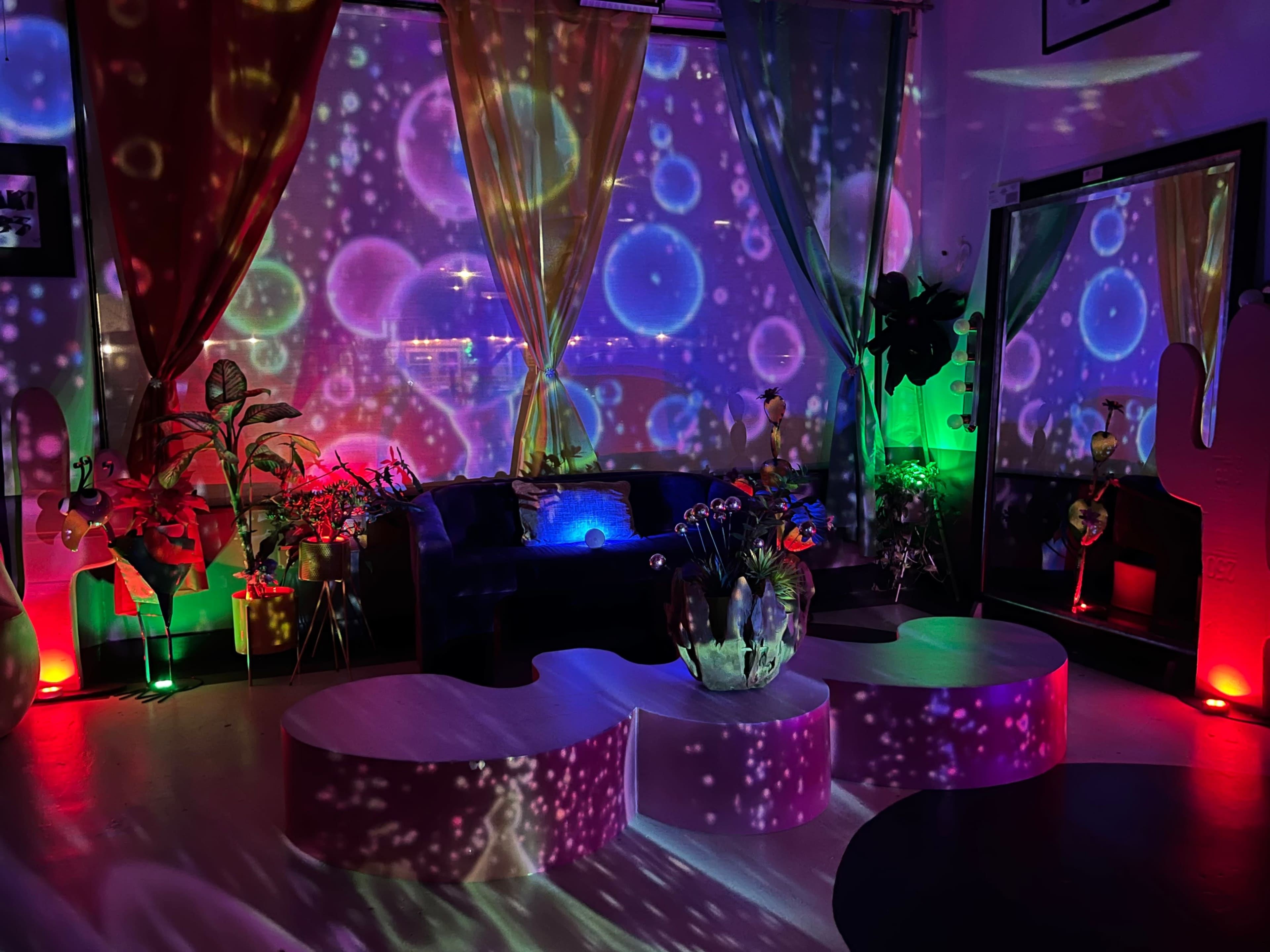 The image depicts a colorful indoor space featuring a dark sofa, two abstract coffee tables, and vibrant light projections creating a bubble-like effect on the walls and floor.
