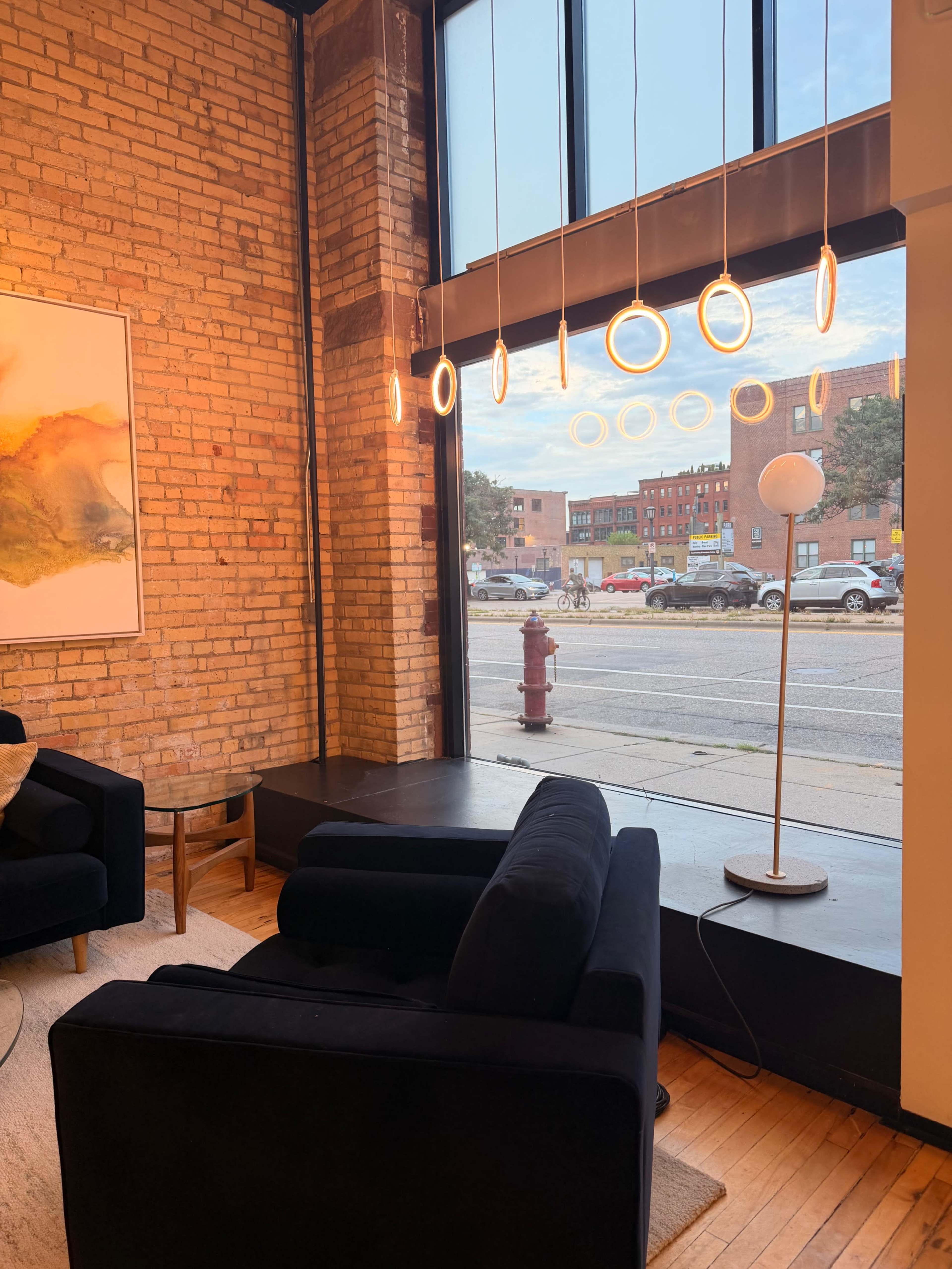 A modern interior features a pair of black chairs and a circular floor lamp near a large window that displays an urban street scene.