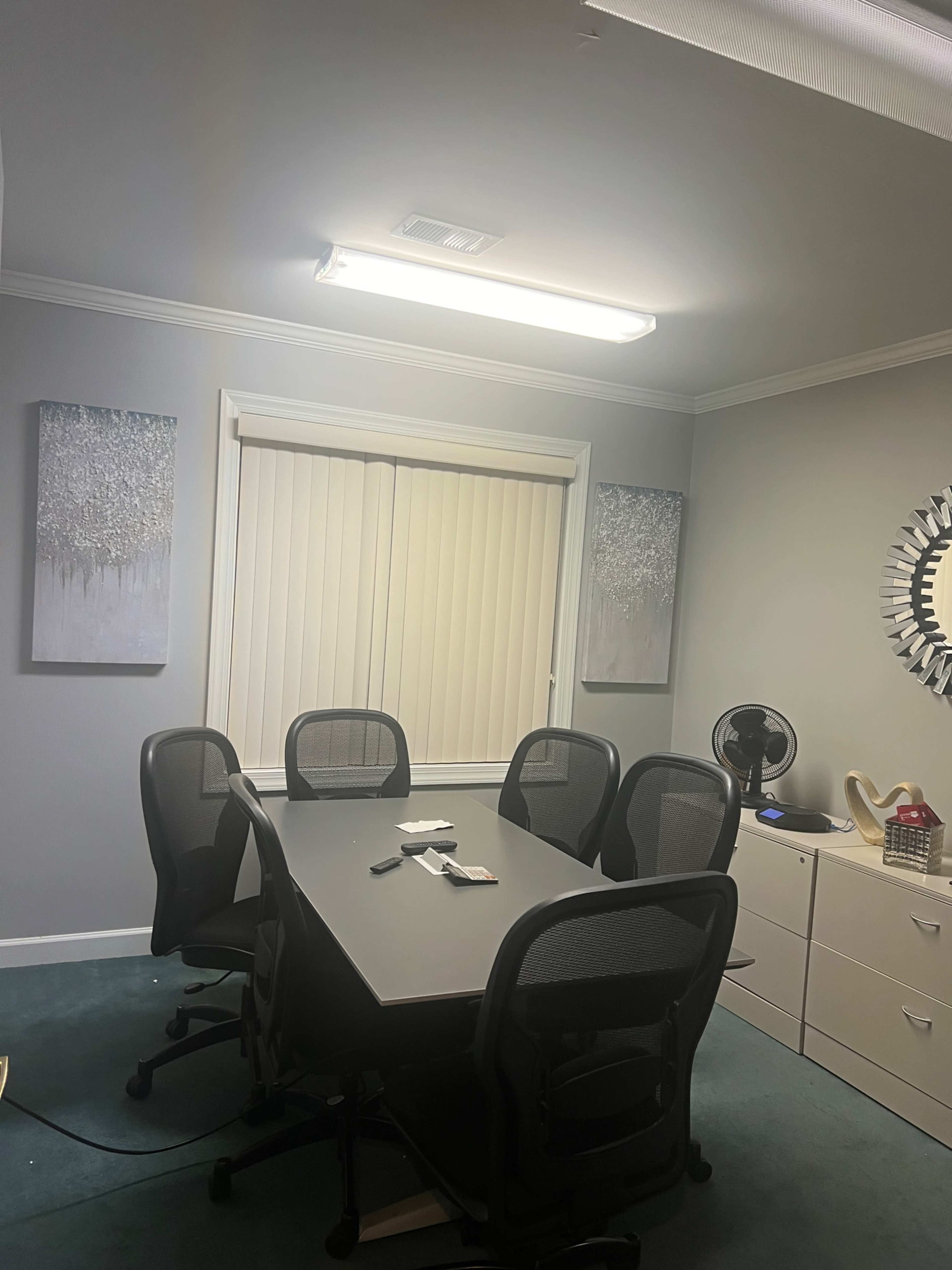 A conference room with a rectangular table, six black mesh chairs, two framed artworks on the walls, and a window covered by horizontal blinds.