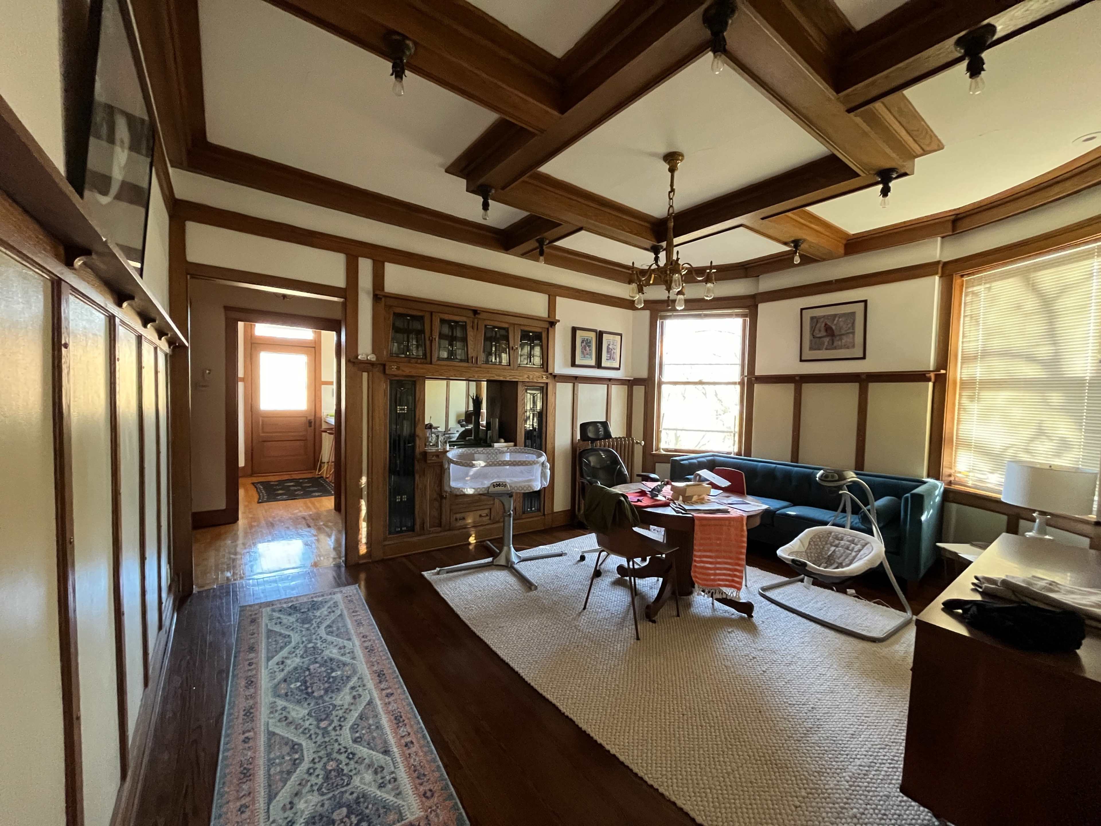 The image shows a room with wooden beam ceilings, featuring a green couch, a dining table with chairs, and an open doorway leading to another area in the house.