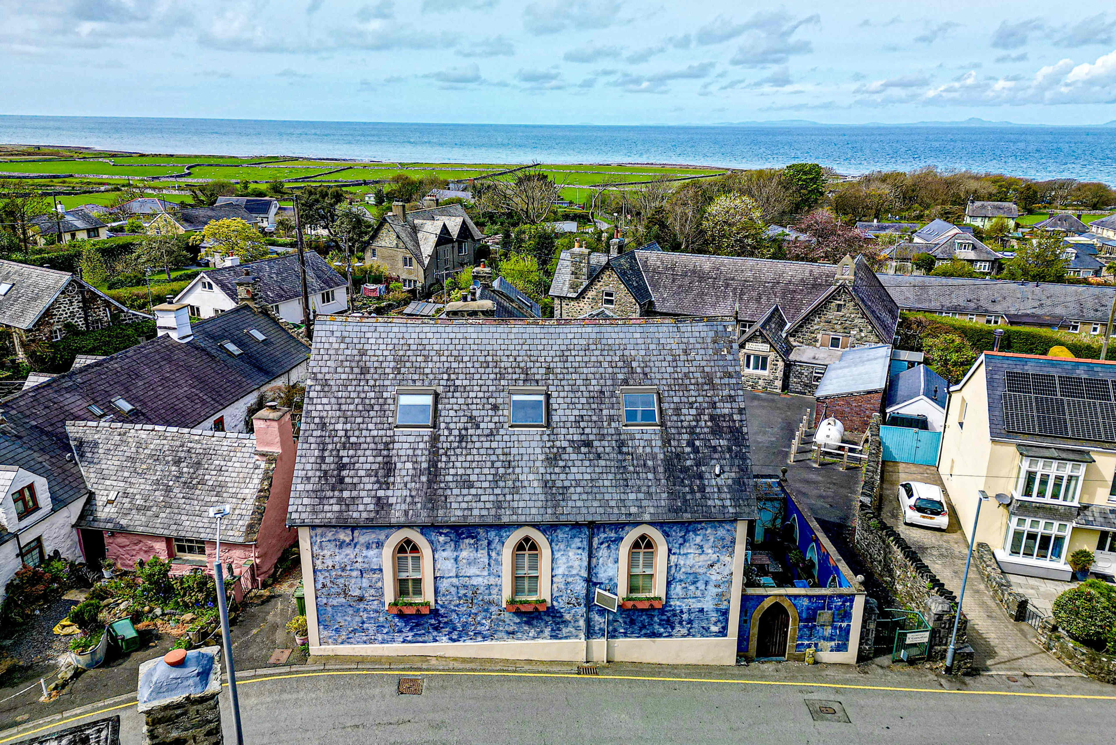 A blue-painted house with gothic-style windows stands at the forefront, surrounded by a quaint village and a view of the ocean in the background.
