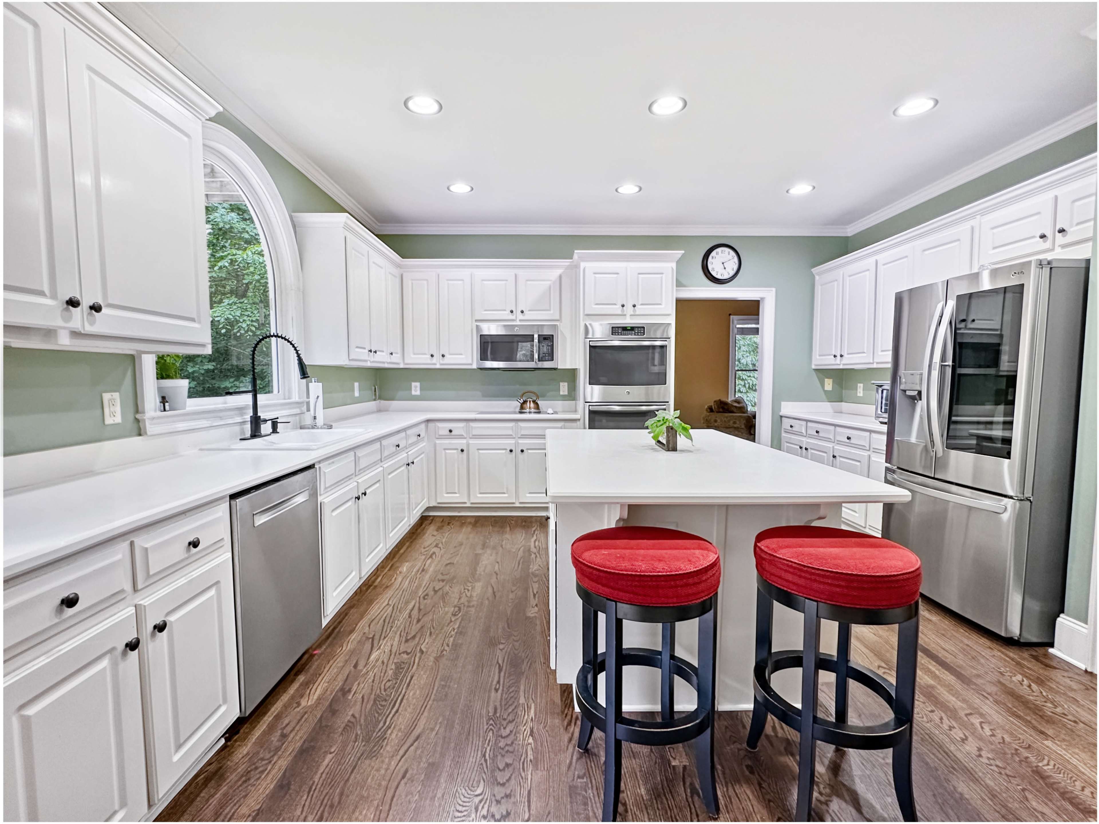 The kitchen features white cabinetry, stainless steel appliances, a central island with red stools, and natural light from a large window.