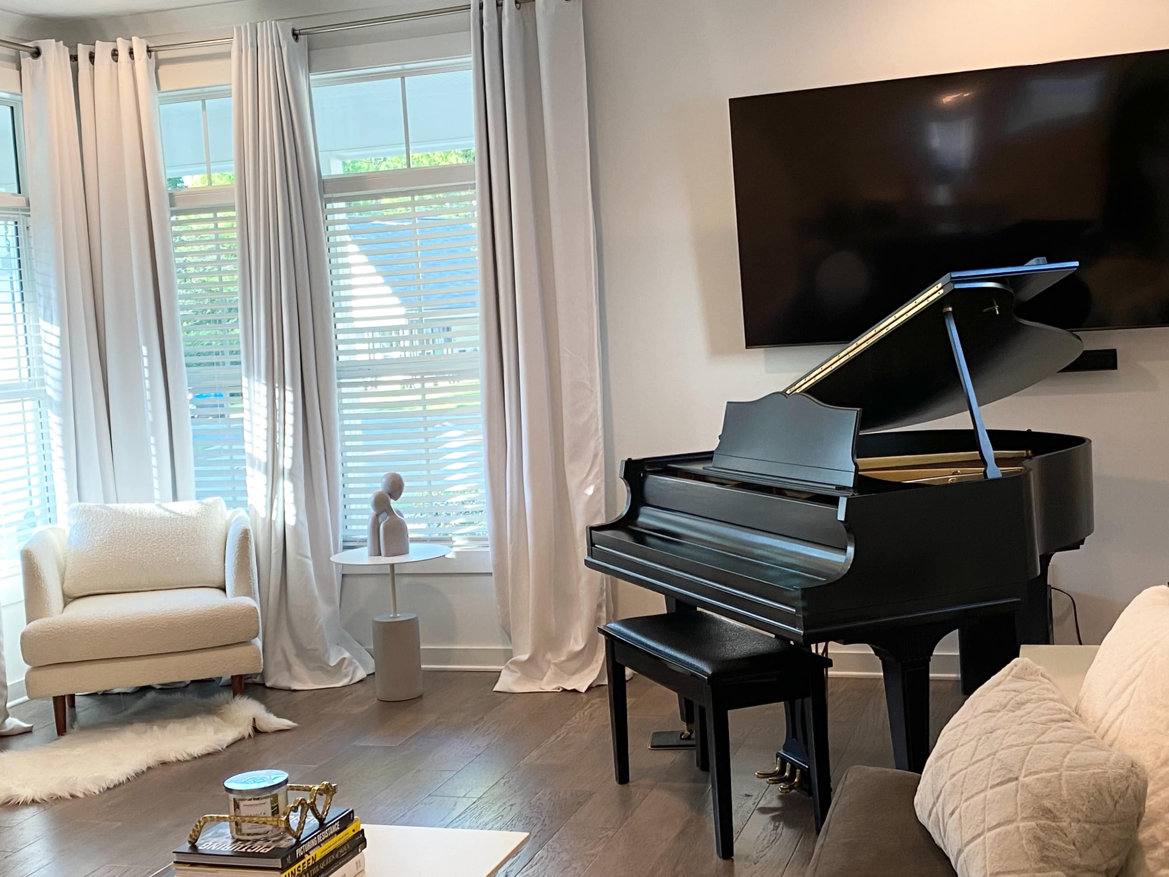 A dark grand piano is positioned beside a modern chair in a bright living room with large windows and a mounted television.