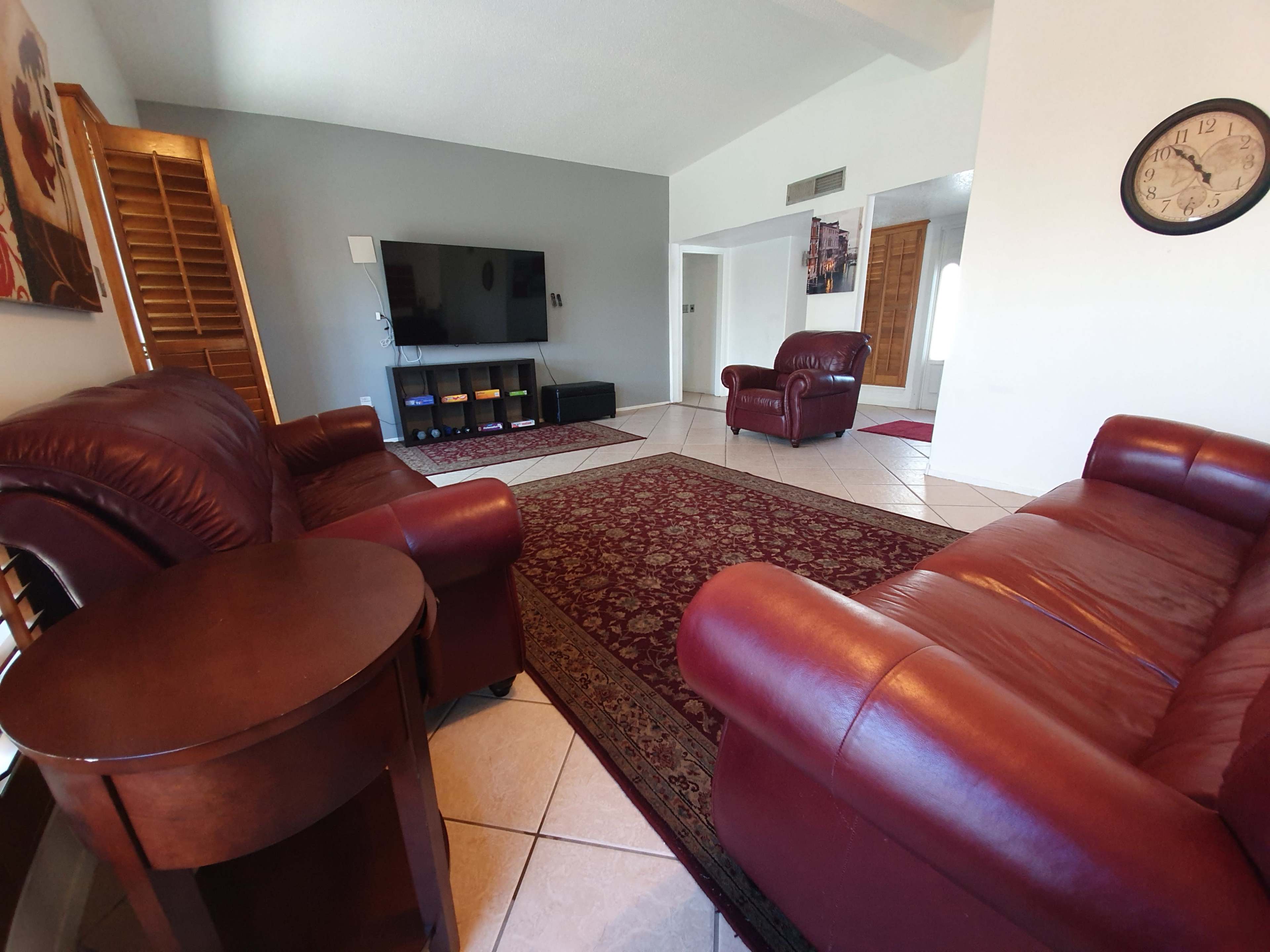 A living room features two red leather chairs and a matching sofa, with a large television on the wall opposite a patterned rug.
