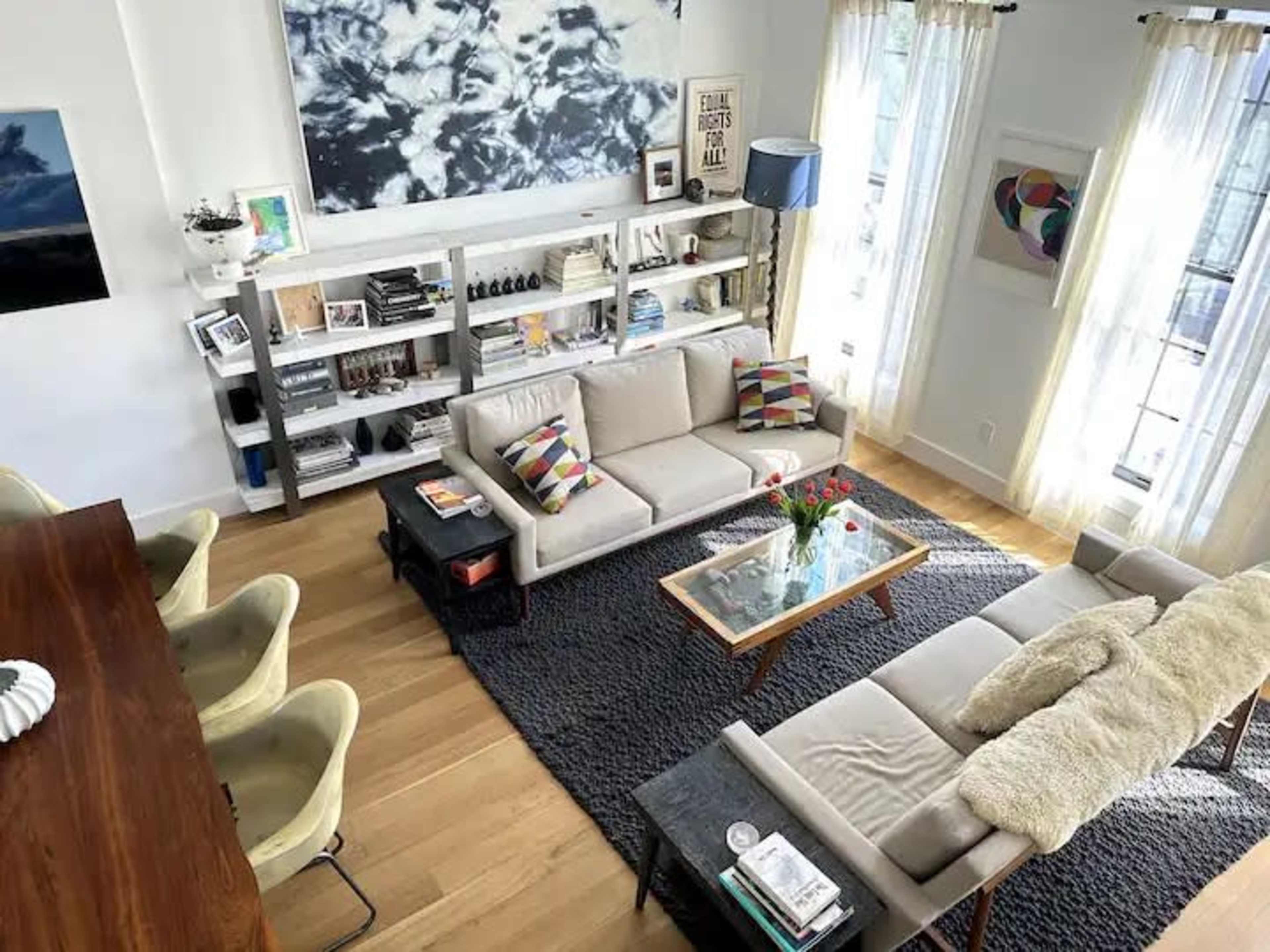 A modern living room features a light-colored sofa with colorful cushions, a glass coffee table, and open shelves filled with books and decorative items, all illuminated by natural light from large windows.