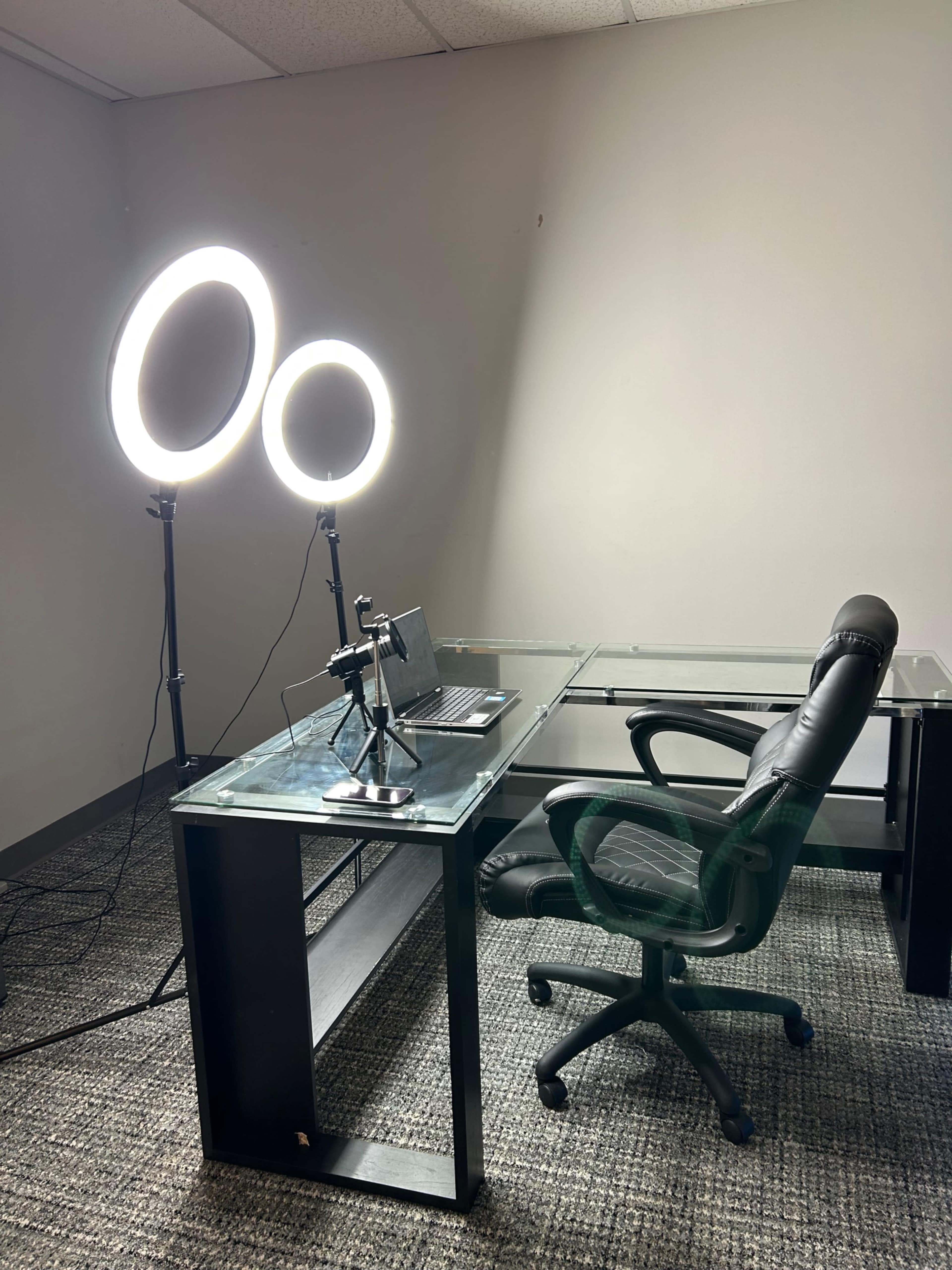 A spacious office setup features a glass desk, a black ergonomic chair, a laptop, and two ring lights positioned behind the desk.