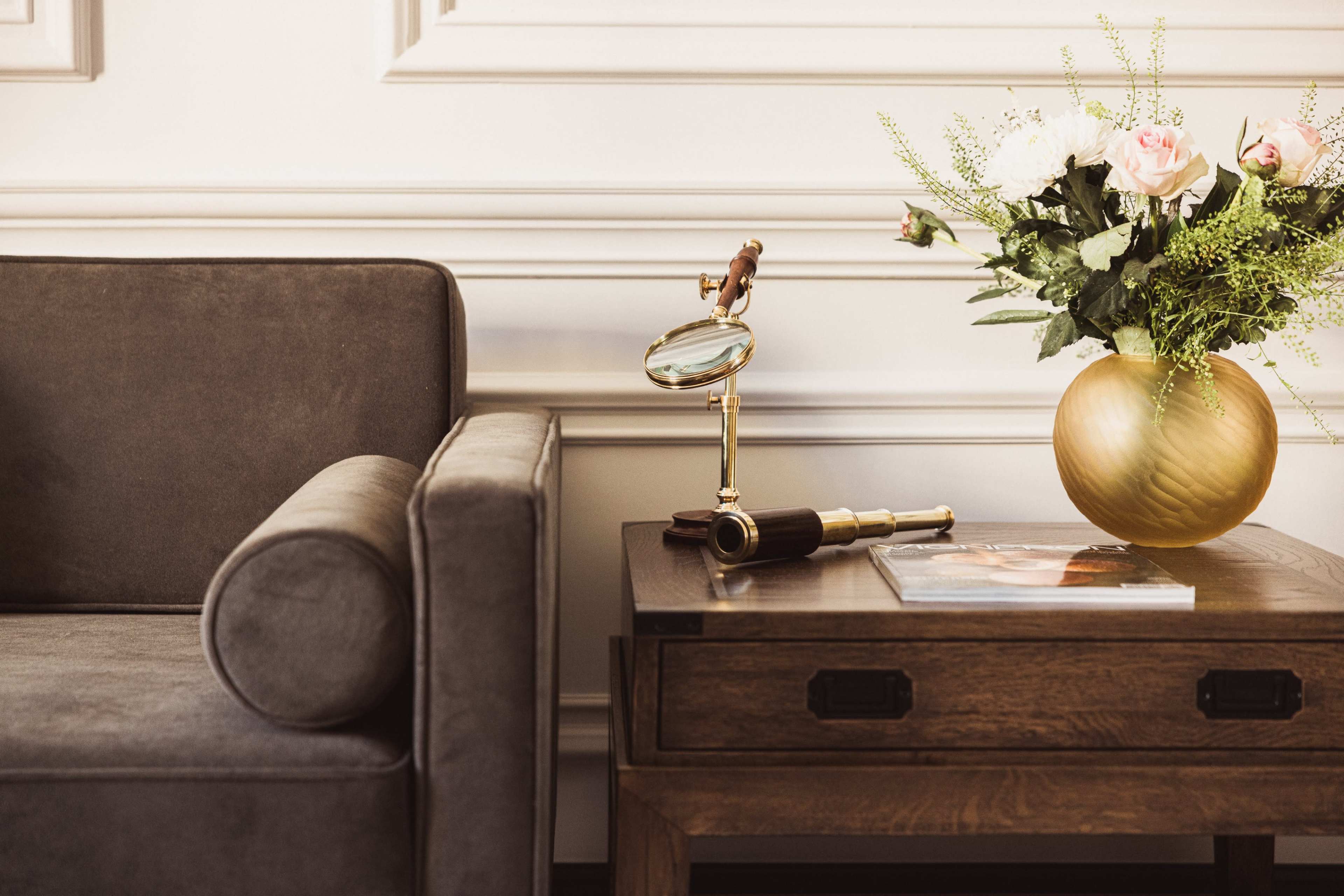 A gray sofa is positioned next to a wooden side table featuring a magnifying glass, a telescope, and a gold vase filled with flowers.
