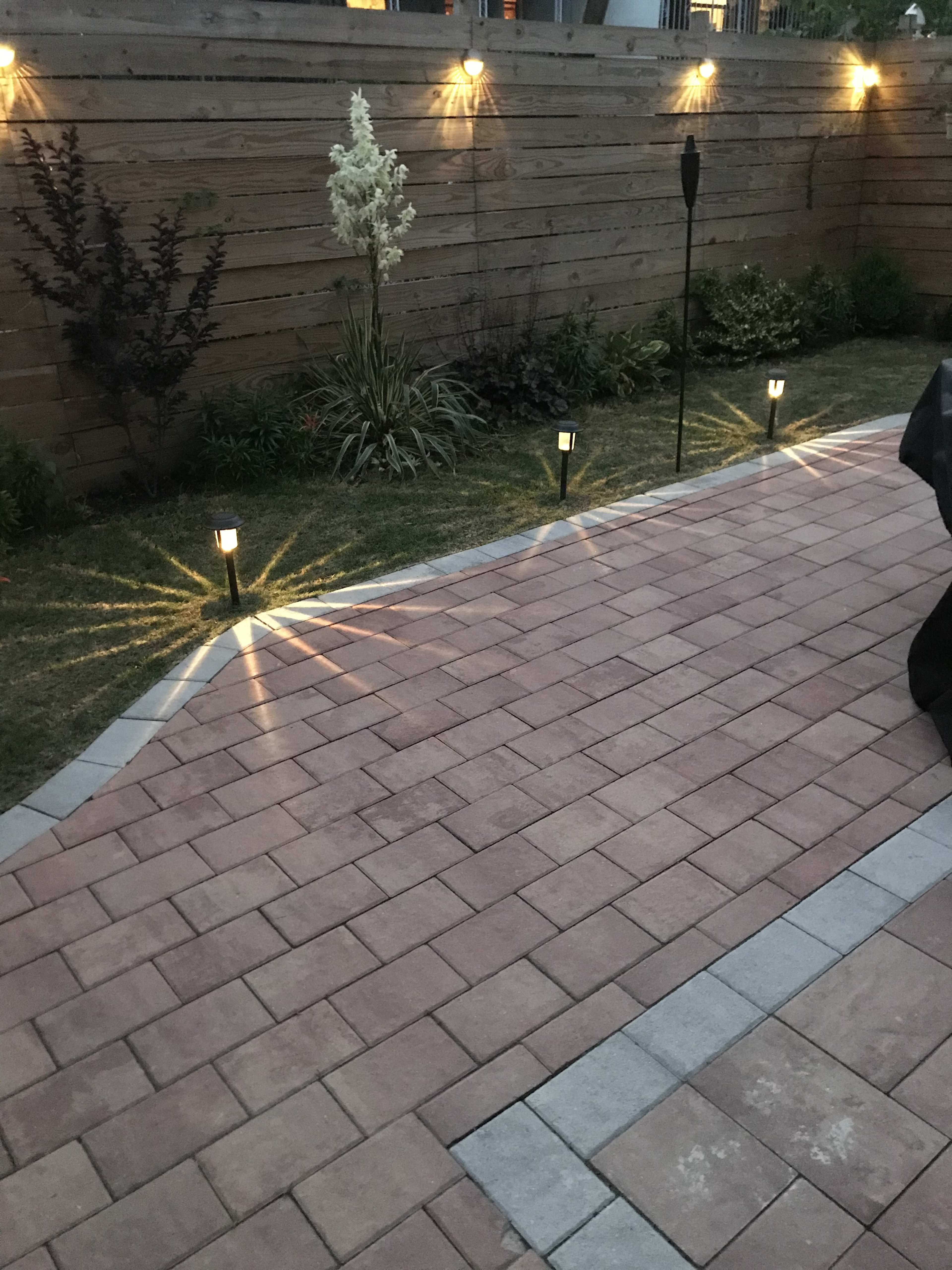 A brick patio illuminated by pathway lights, with a wooden fence and landscaped greenery in the background.