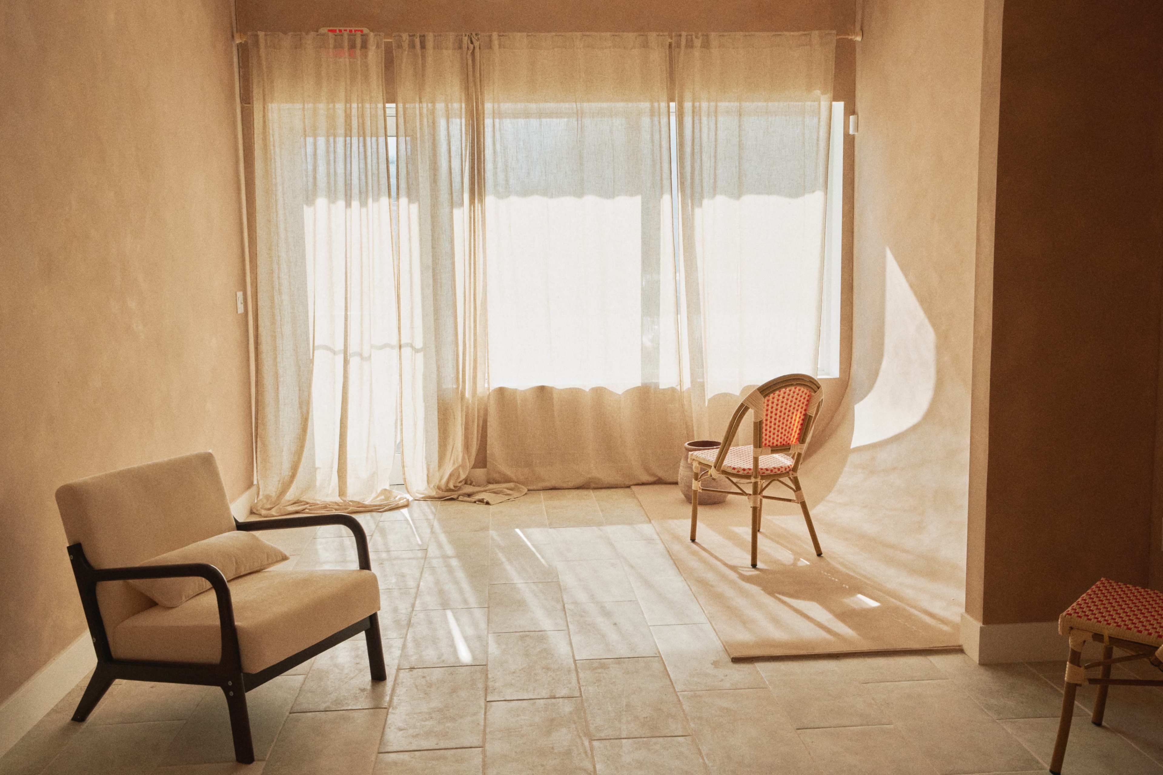 The image shows a light-filled room with a pair of chairs, a window covered by sheer curtains, and a soft area rug on the tiled floor.