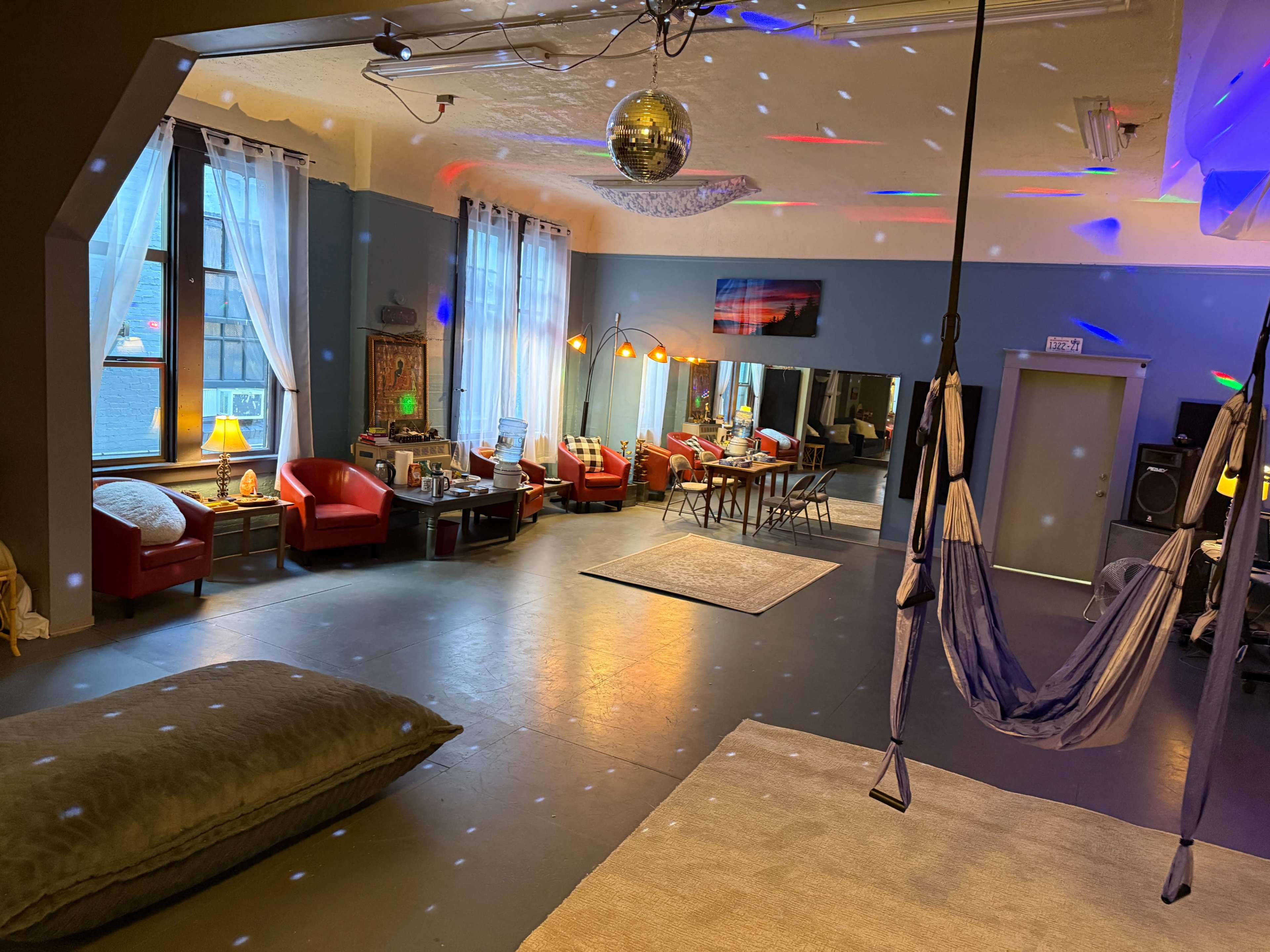 The image shows a spacious room with colorful lights reflecting on the floor, equipped with red chairs, a table, and a hammock, suggesting a social or lounge area.