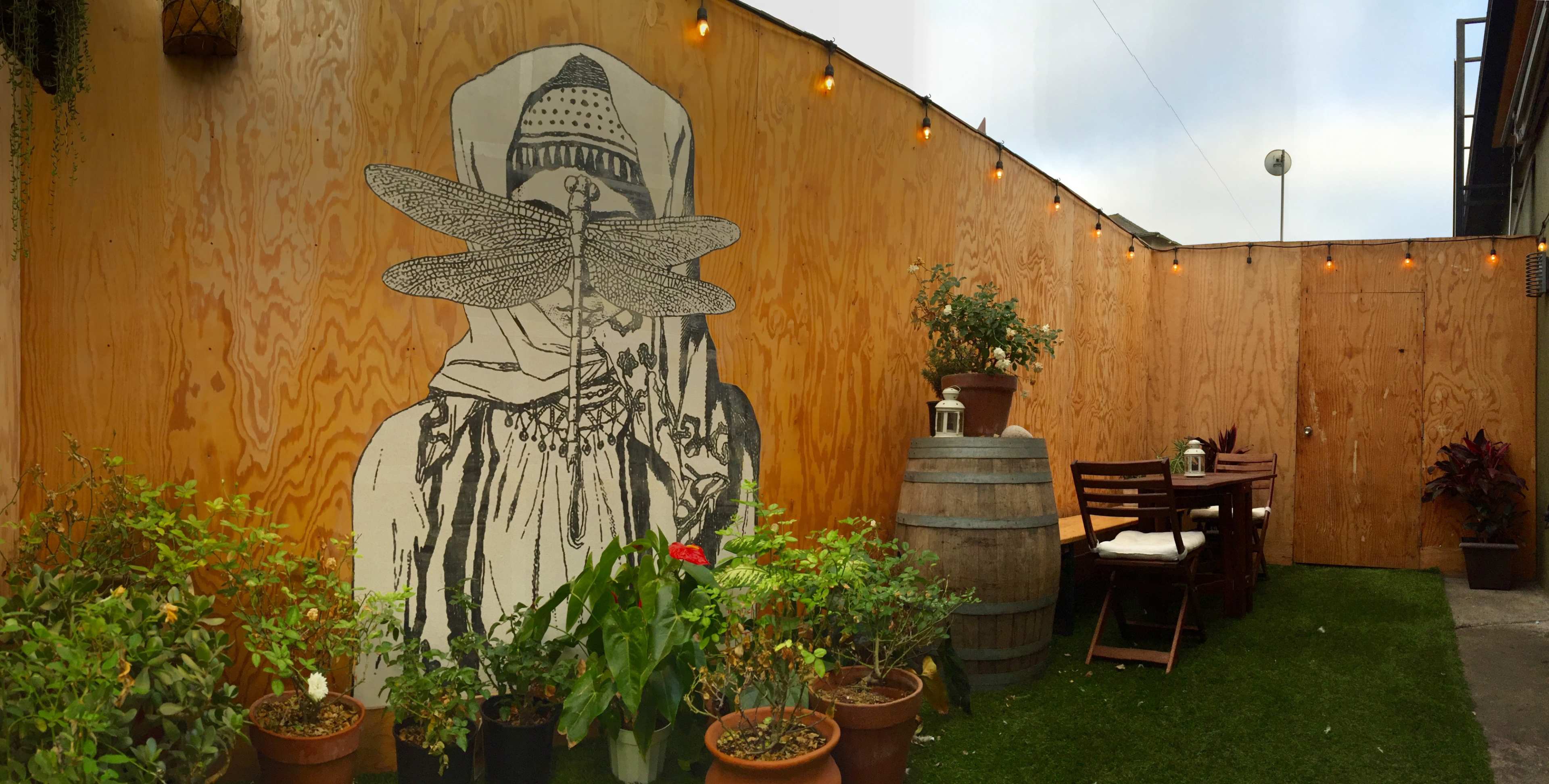 A mural of a person with a hat and dragonflies is painted on a wooden wall in a garden area with potted plants and outdoor seating.