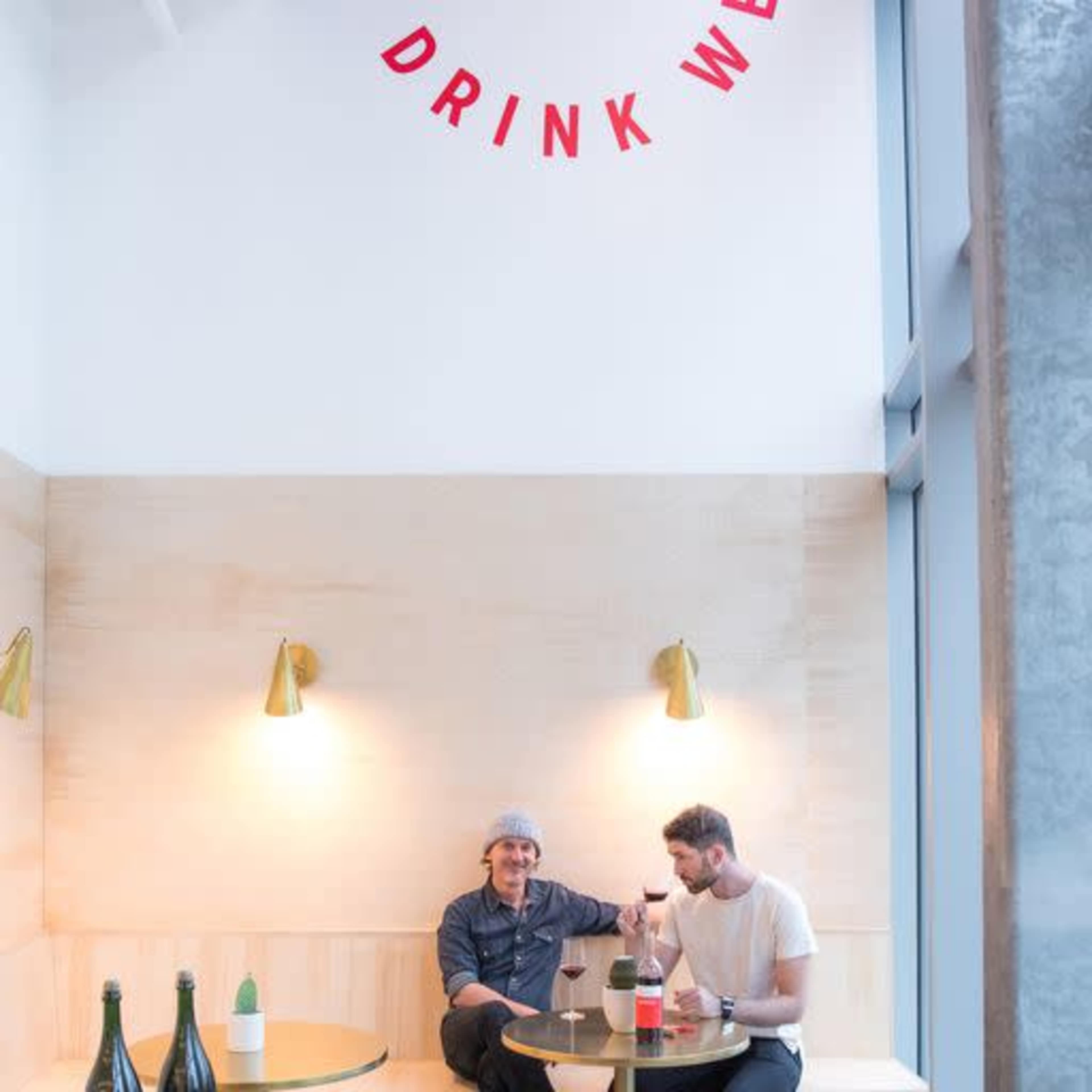 Two men sit at a small table in a bright interior, enjoying wine bottles and glasses under modern lighting.