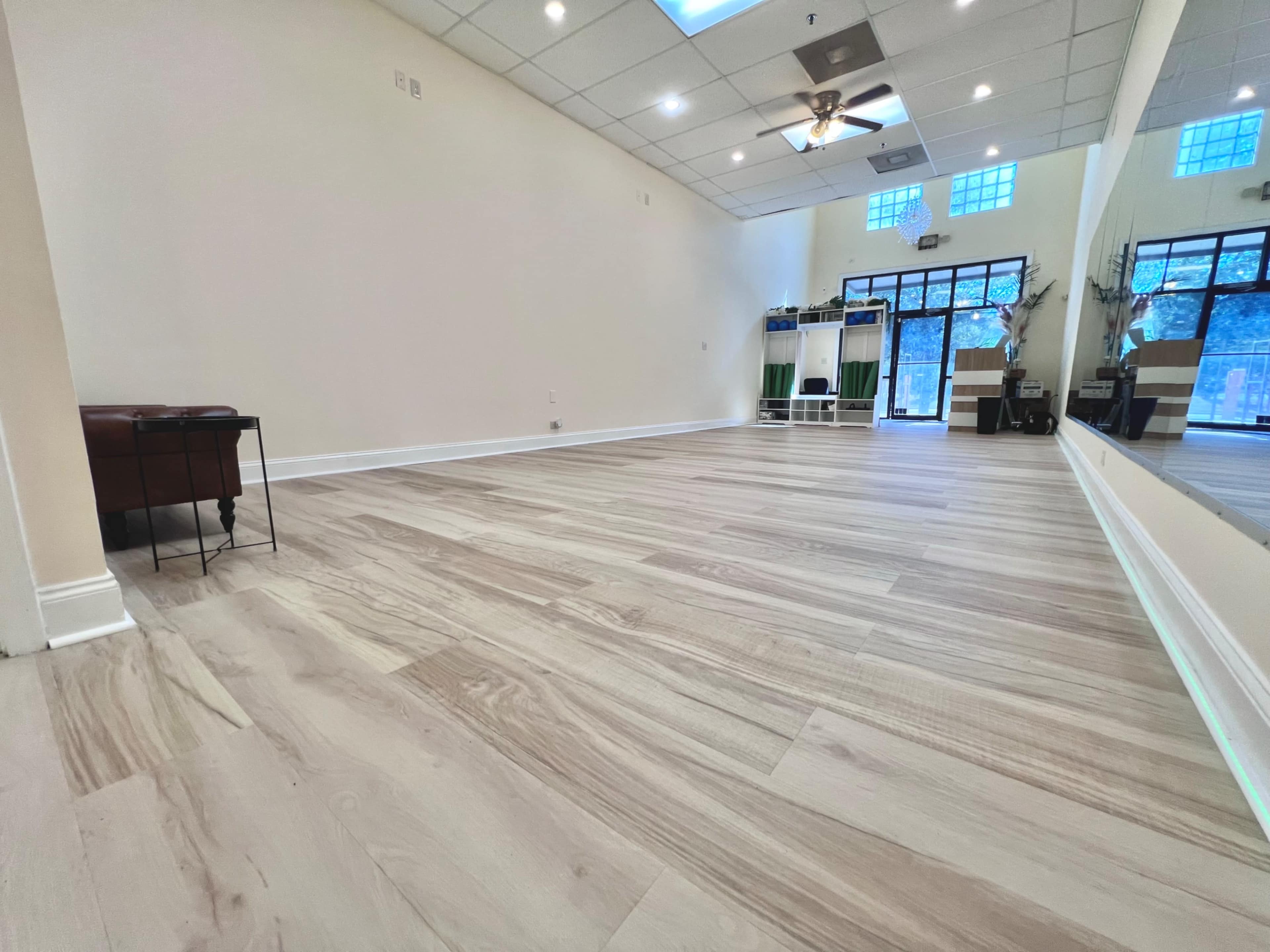 The image shows a spacious, empty room with wooden flooring, white walls, and large windows allowing natural light to enter.