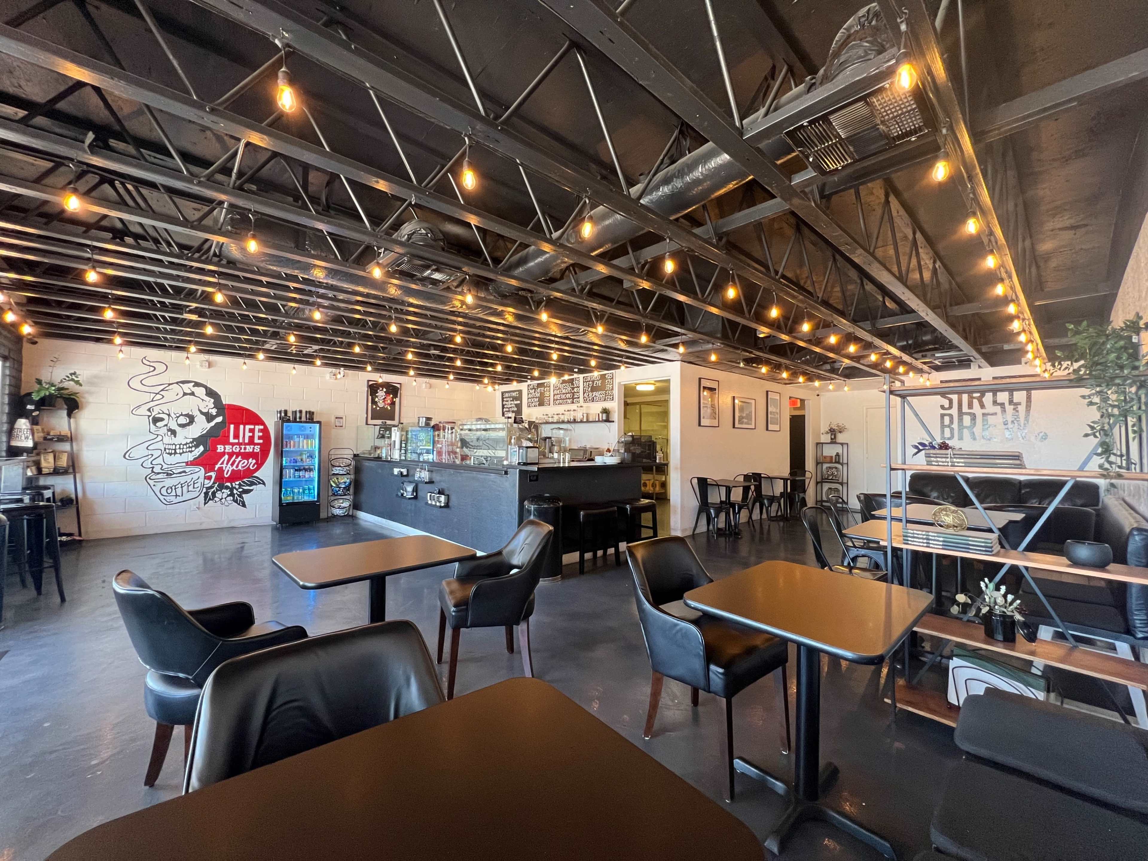 A well-lit cafe interior featuring black tables and chairs, exposed beams, string lights, and a counter with a menu and display cases.