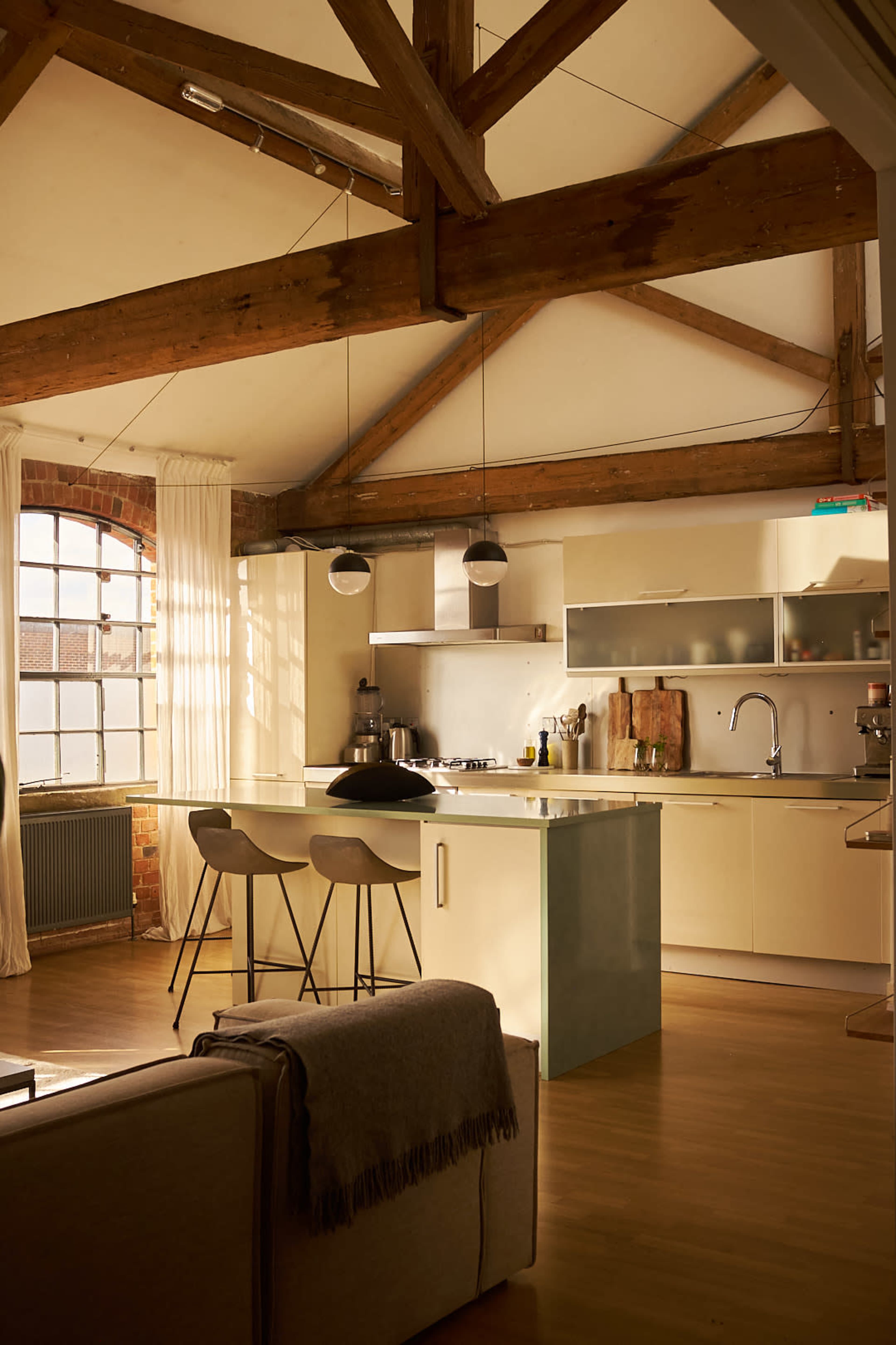 The image shows a modern kitchen with wooden beams, featuring a gas stove, sleek cabinetry, and two bar stools.