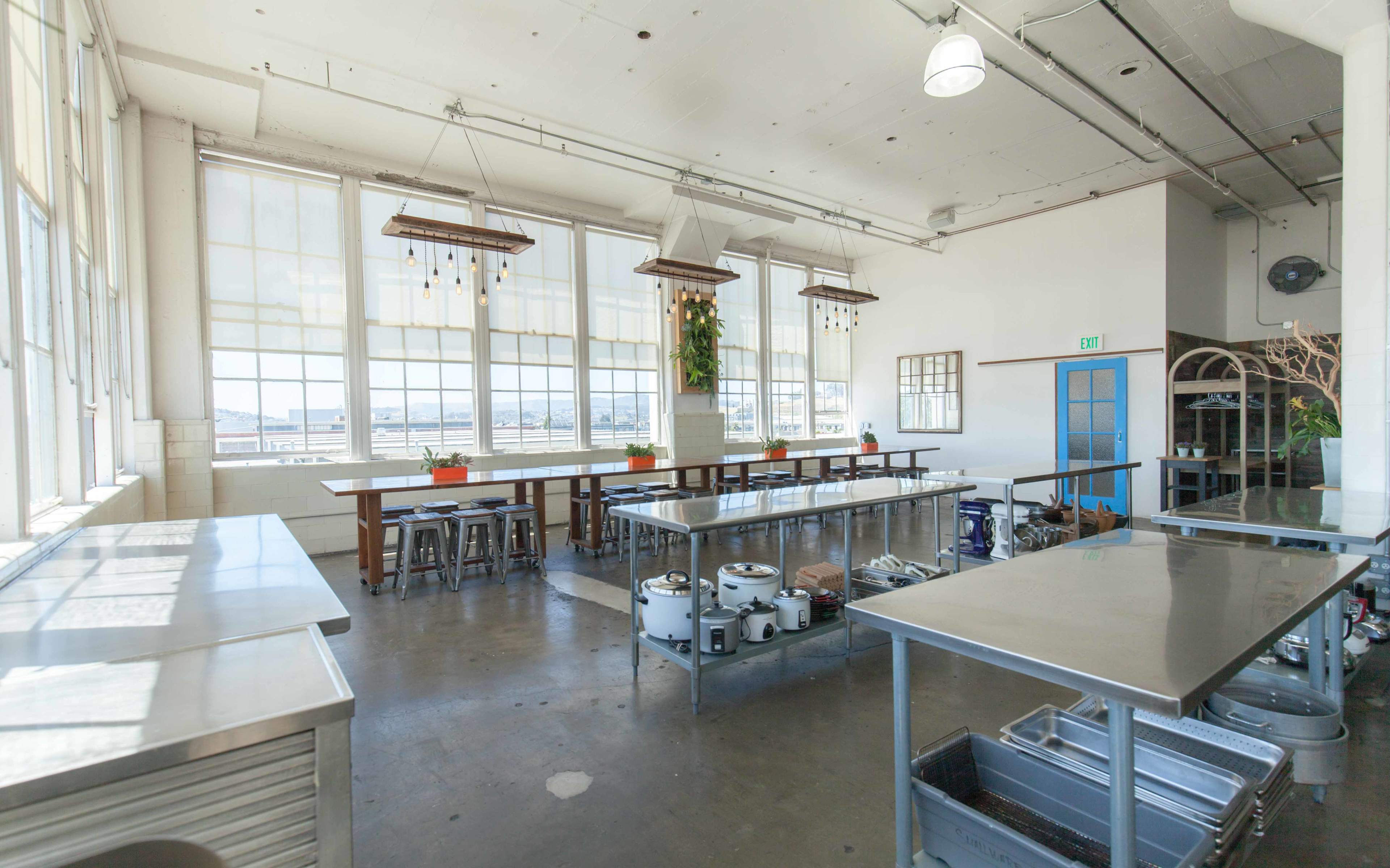 A spacious kitchen with large windows, stainless steel worktables, and overhead lighting.