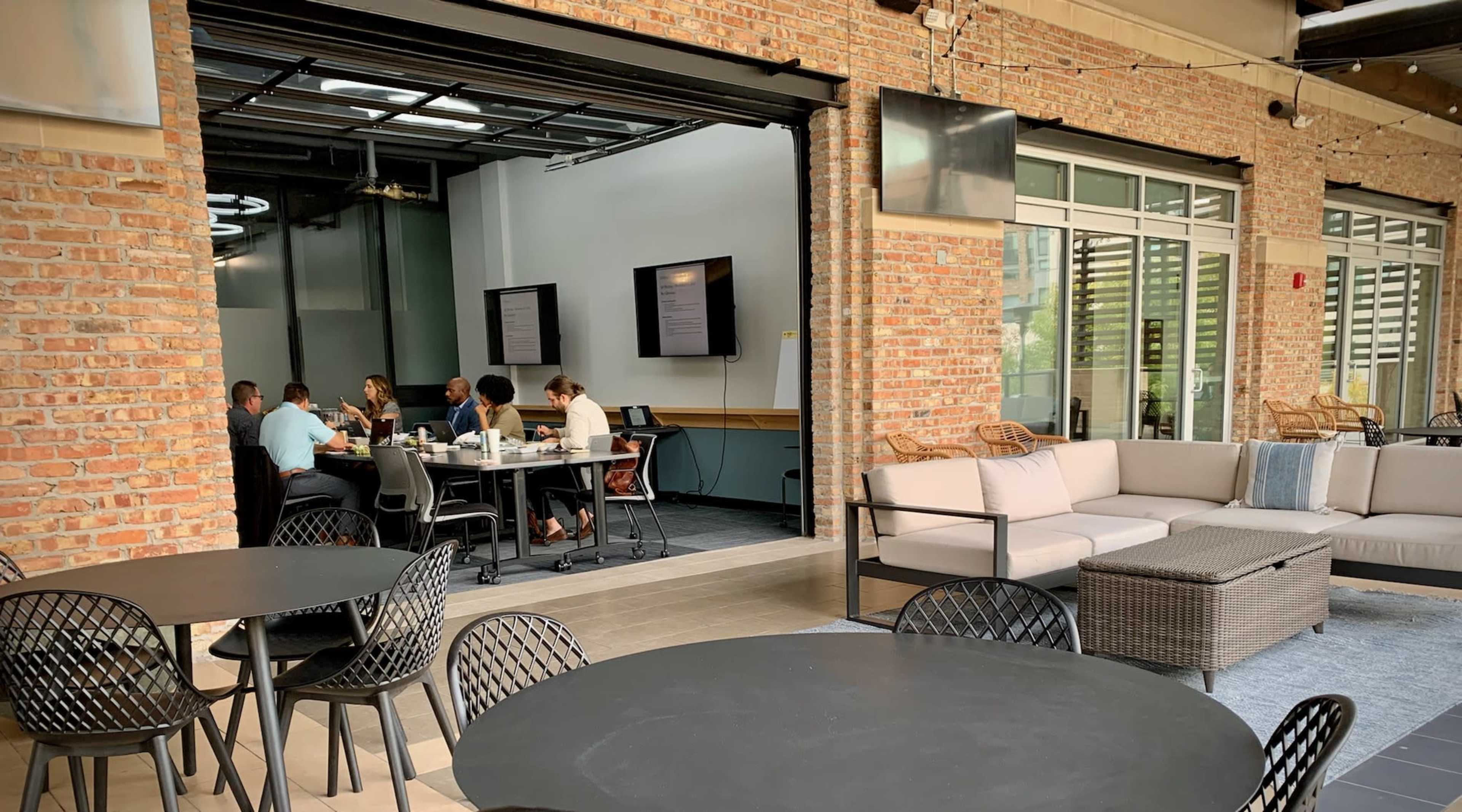 A group of people is seated around a table in a meeting room, while others relax on sofas in an open office space with brick walls.