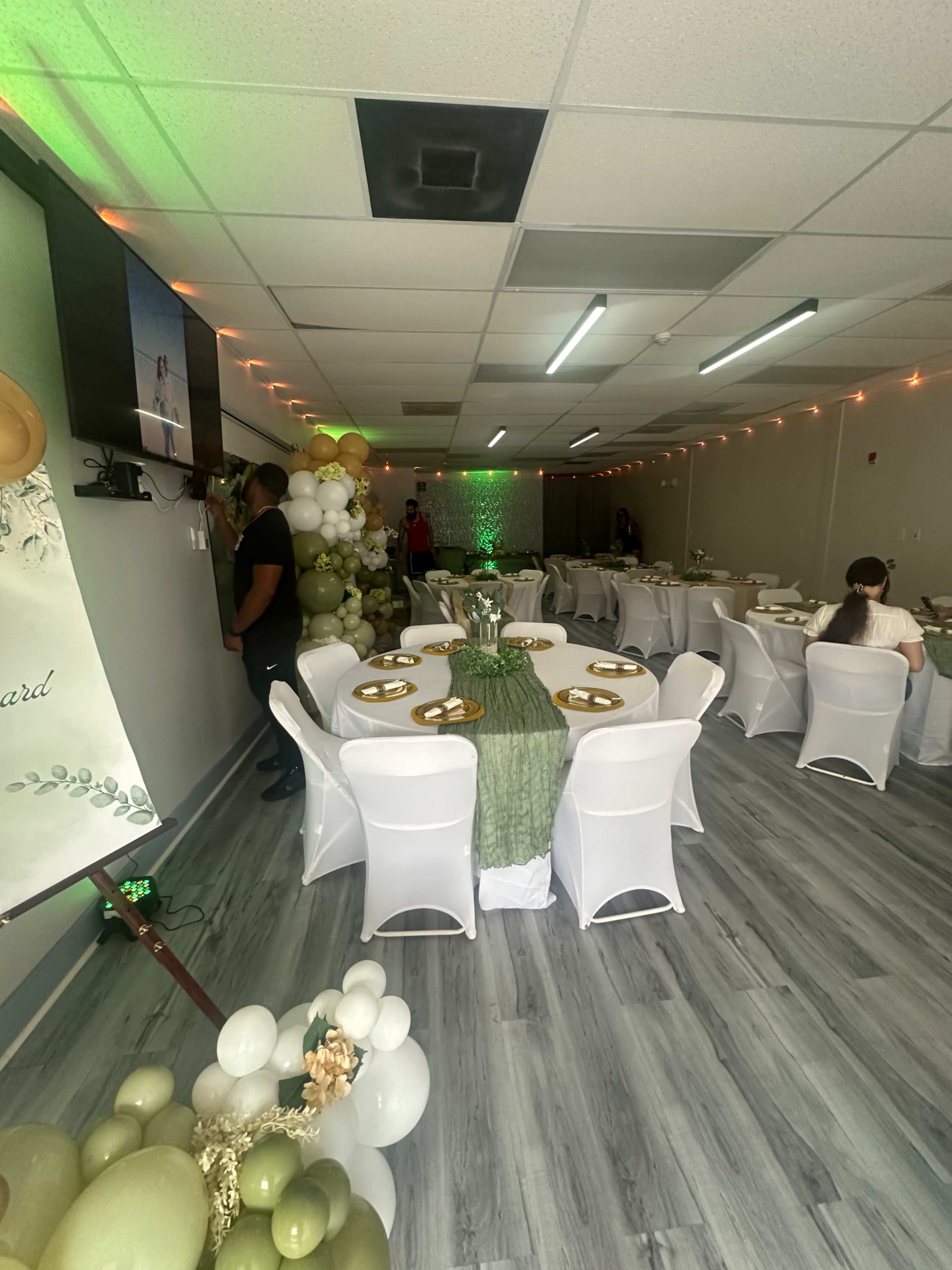 The image shows a decorated event space featuring tables set with white chairs and green tablecloths, adorned with balloons and a television displaying a presentation.