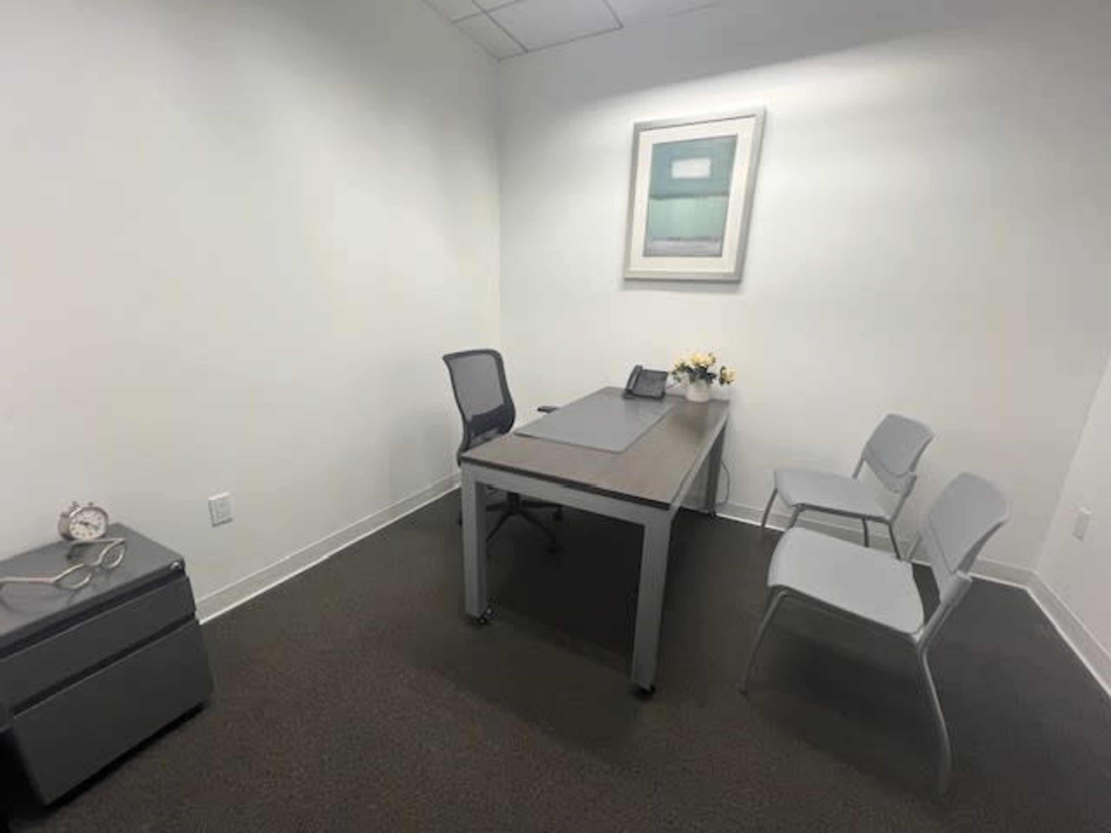 A small office features a desk with a chair, two empty chairs, and a wall art piece above the desk.