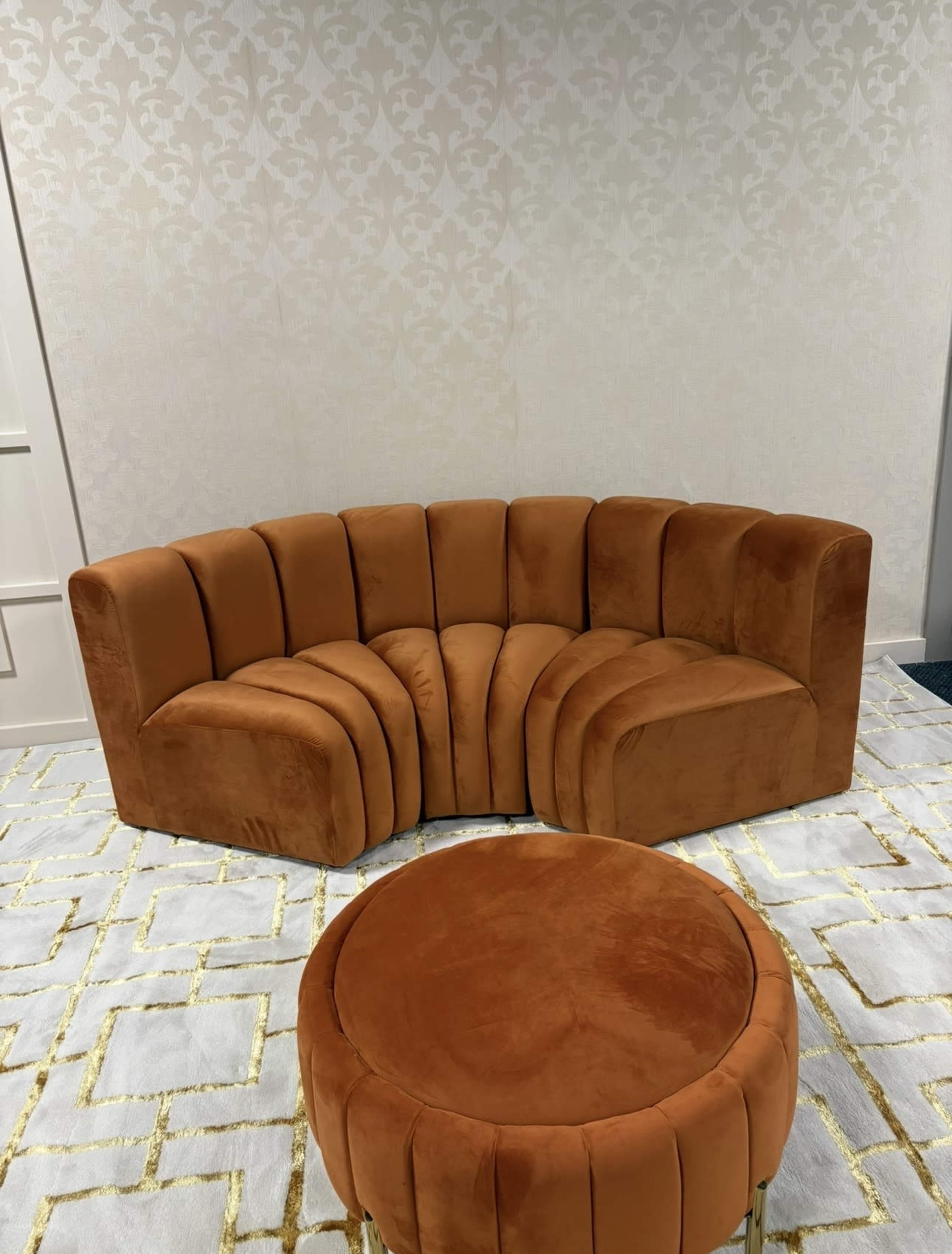 The image shows a curved orange velvet sofa accompanied by a matching round ottoman, set against a light-colored patterned wall.