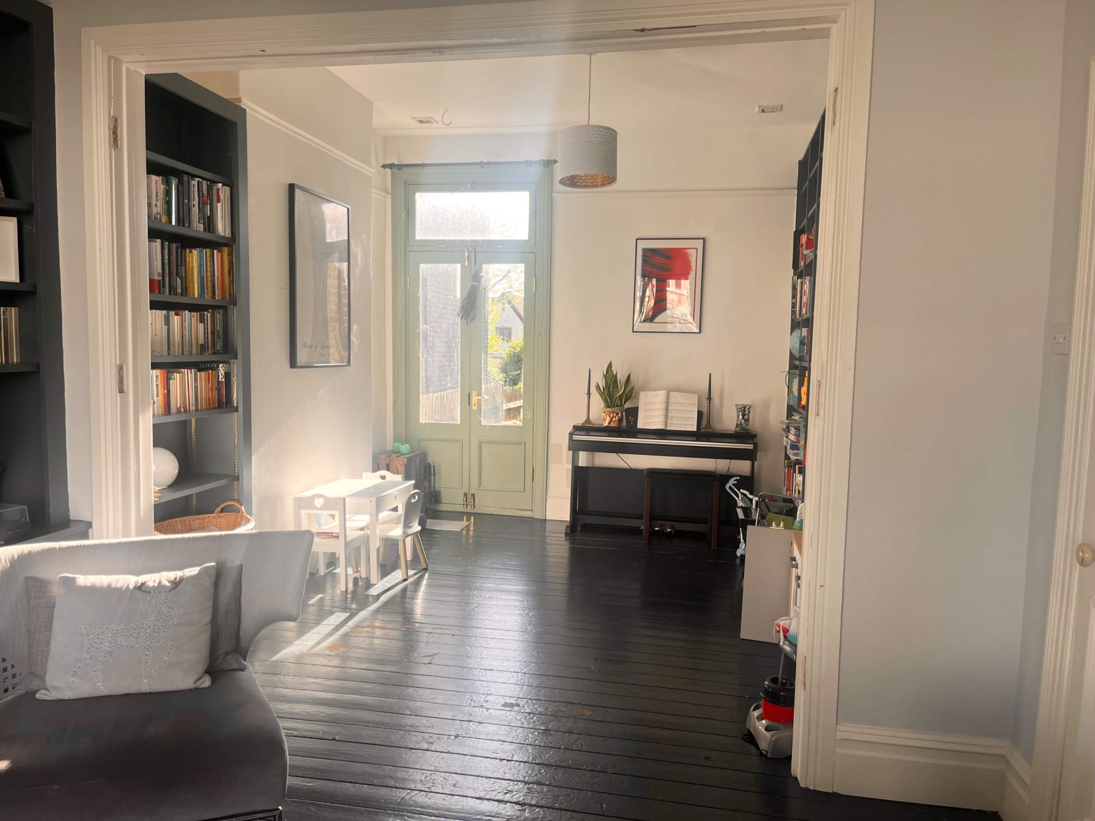 A well-lit room featuring a doorway leading to a space with a green door, bookshelves, a piano, and a small table with chairs.