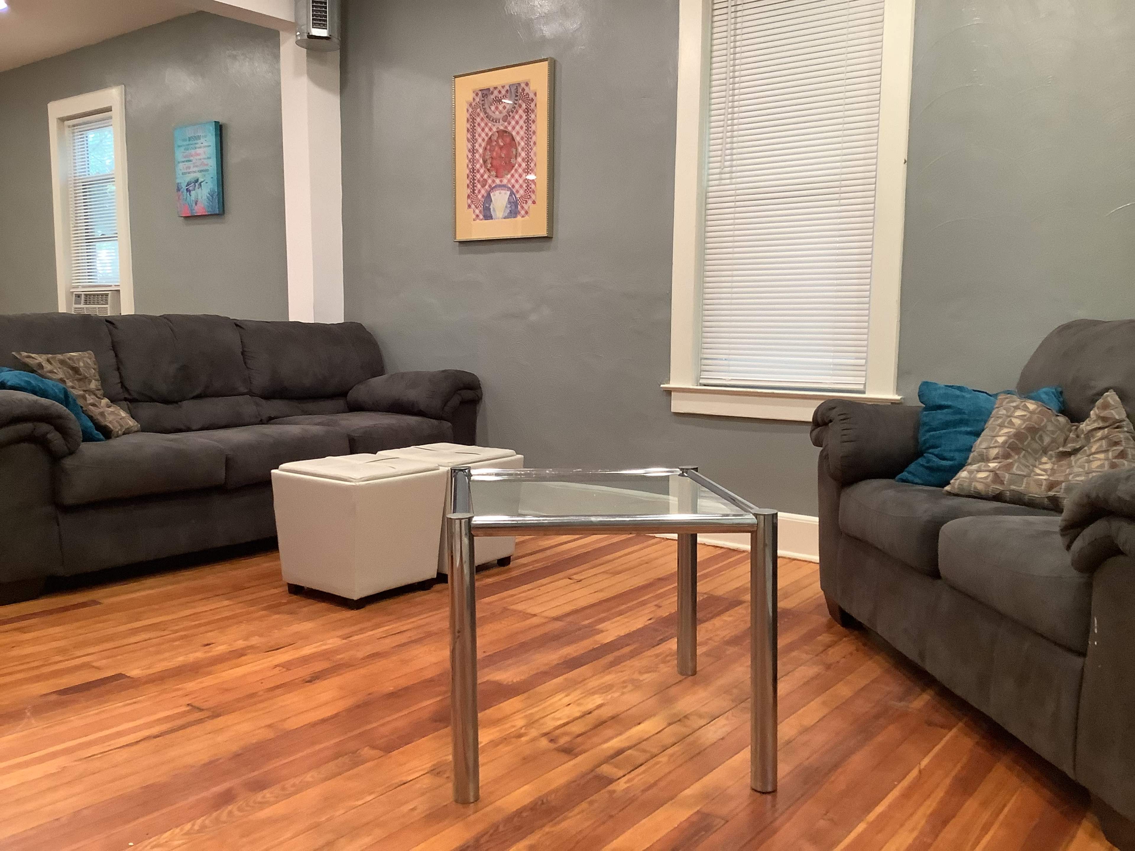 The living room features two dark couches, a glass coffee table with metallic legs, and a white ottoman on wooden flooring.