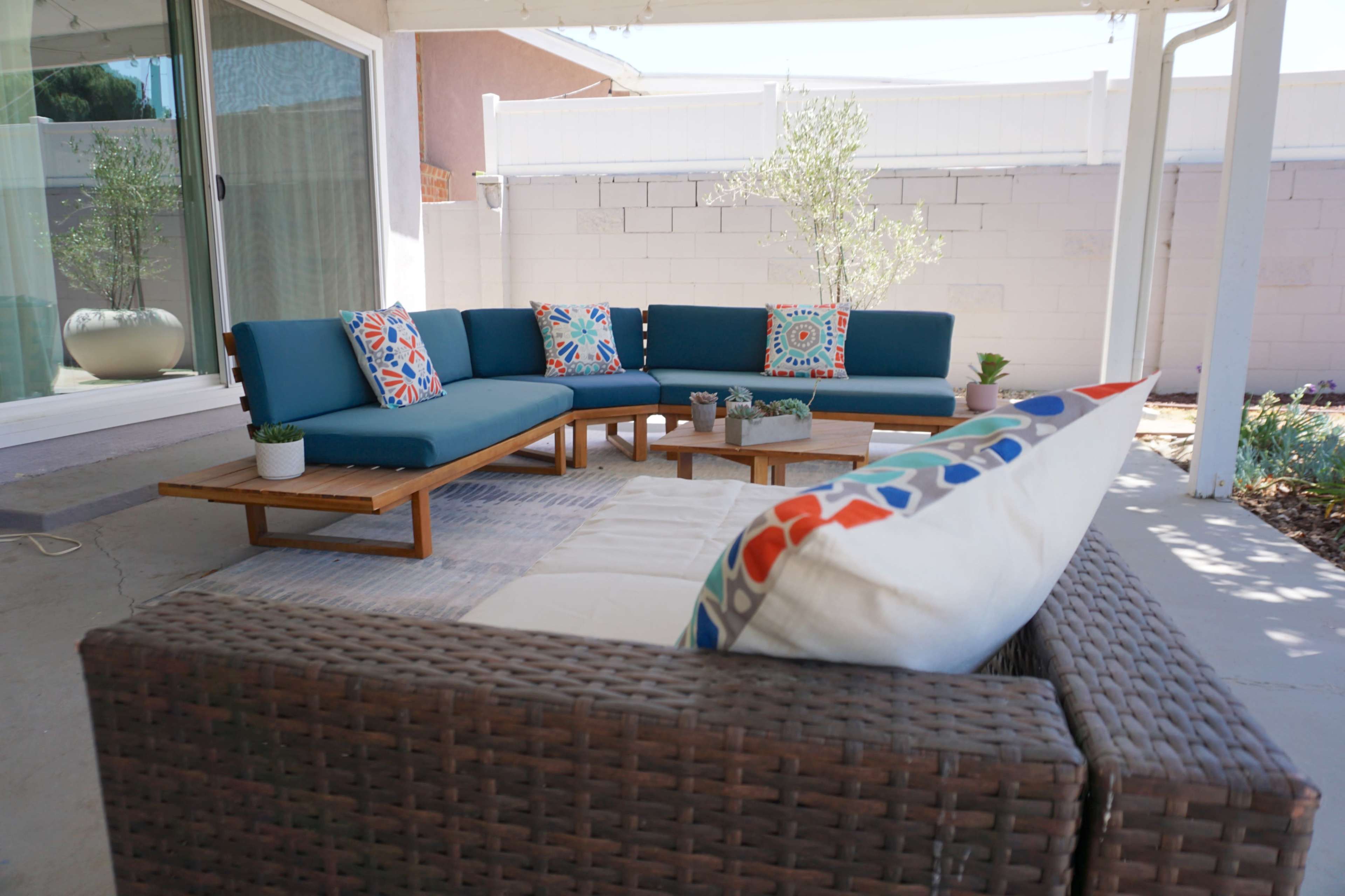 A comfortable outdoor seating area features a blue sectional sofa with colorful cushions and a wooden coffee table, surrounded by a light-colored concrete patio.