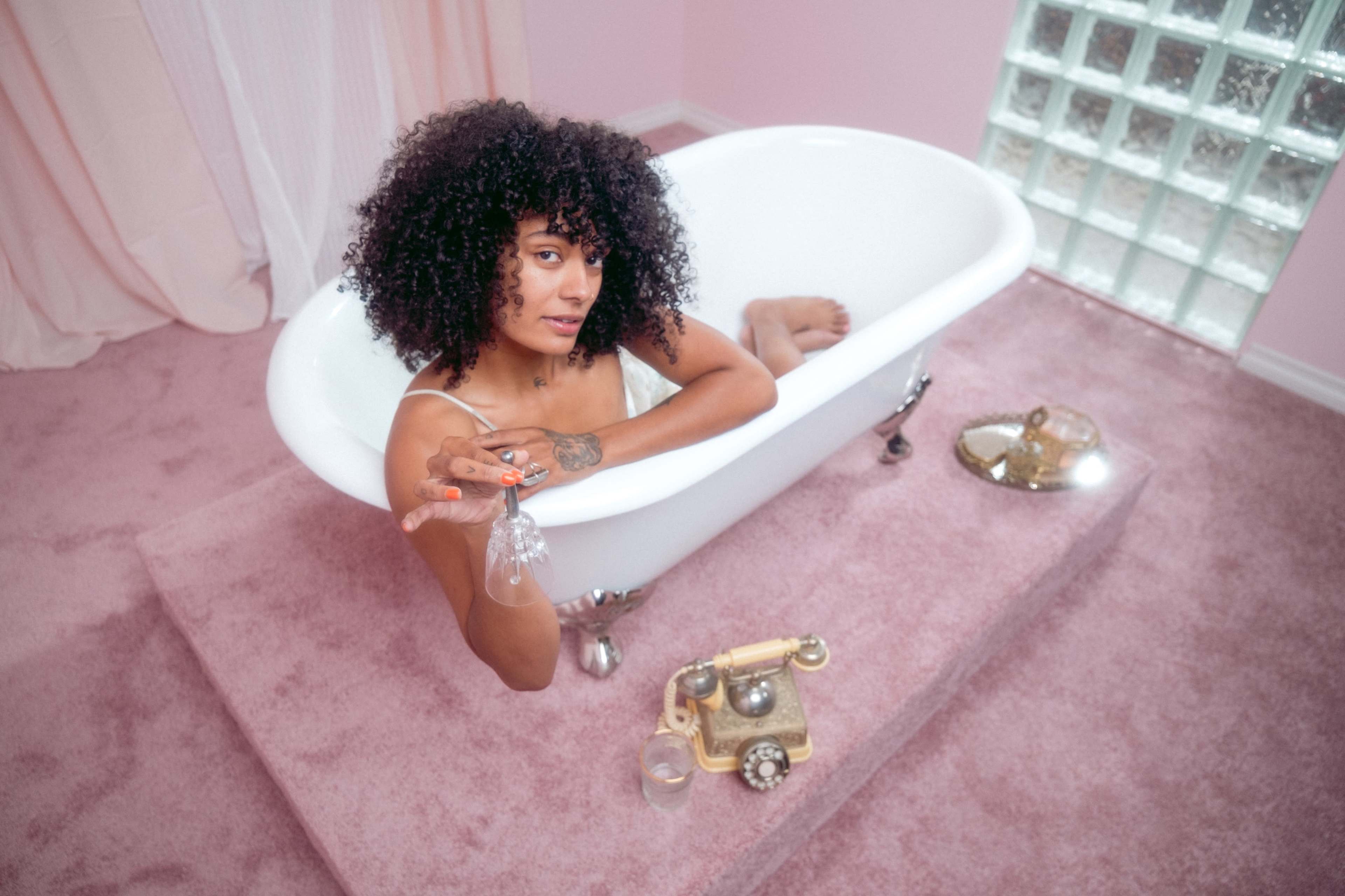 A person with curly hair sits in a white bathtub, surrounded by pink walls and carpeting, holding a glass while looking toward the camera.