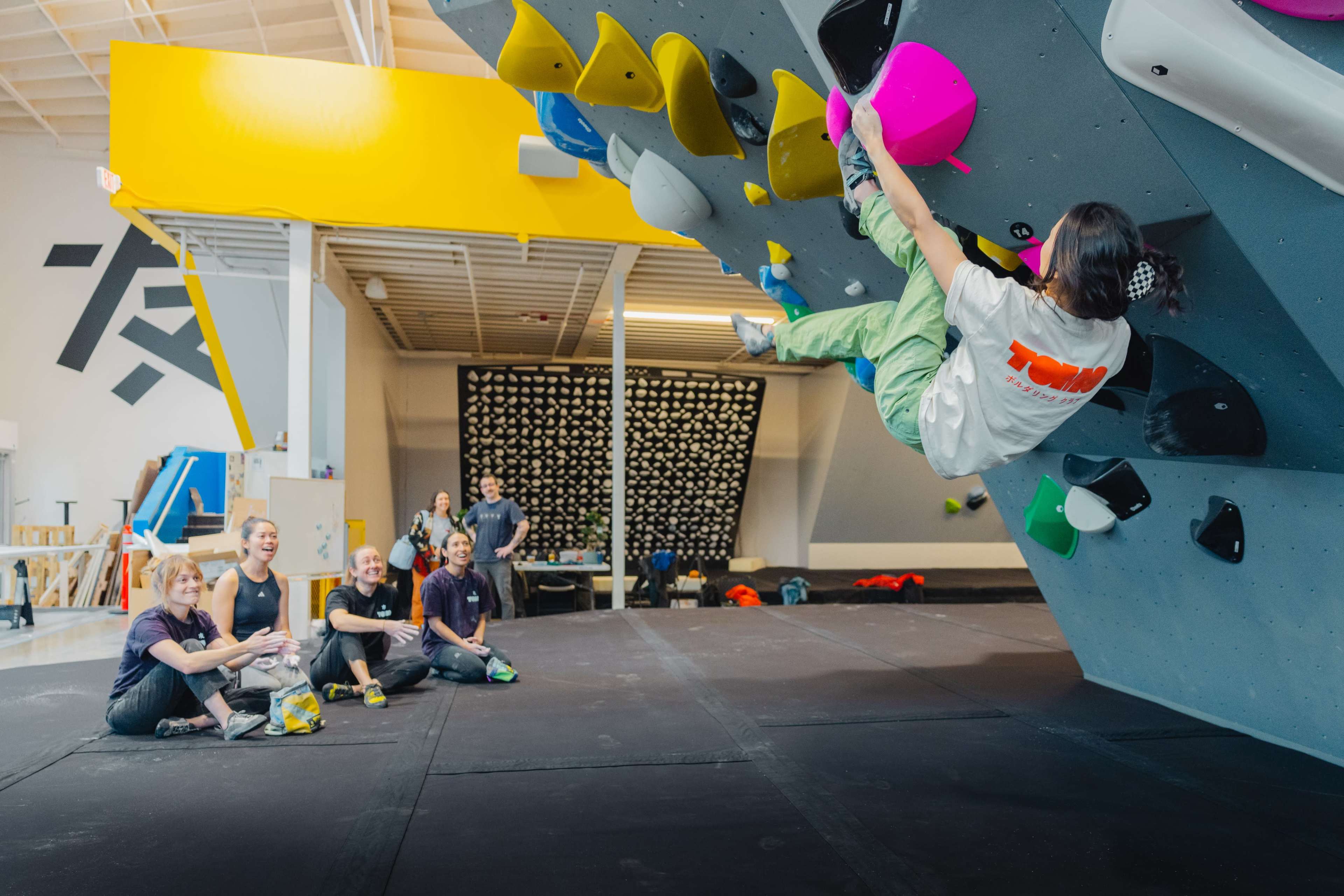 A person climbs on a bouldering wall while a group of five spectators watch from the floor.