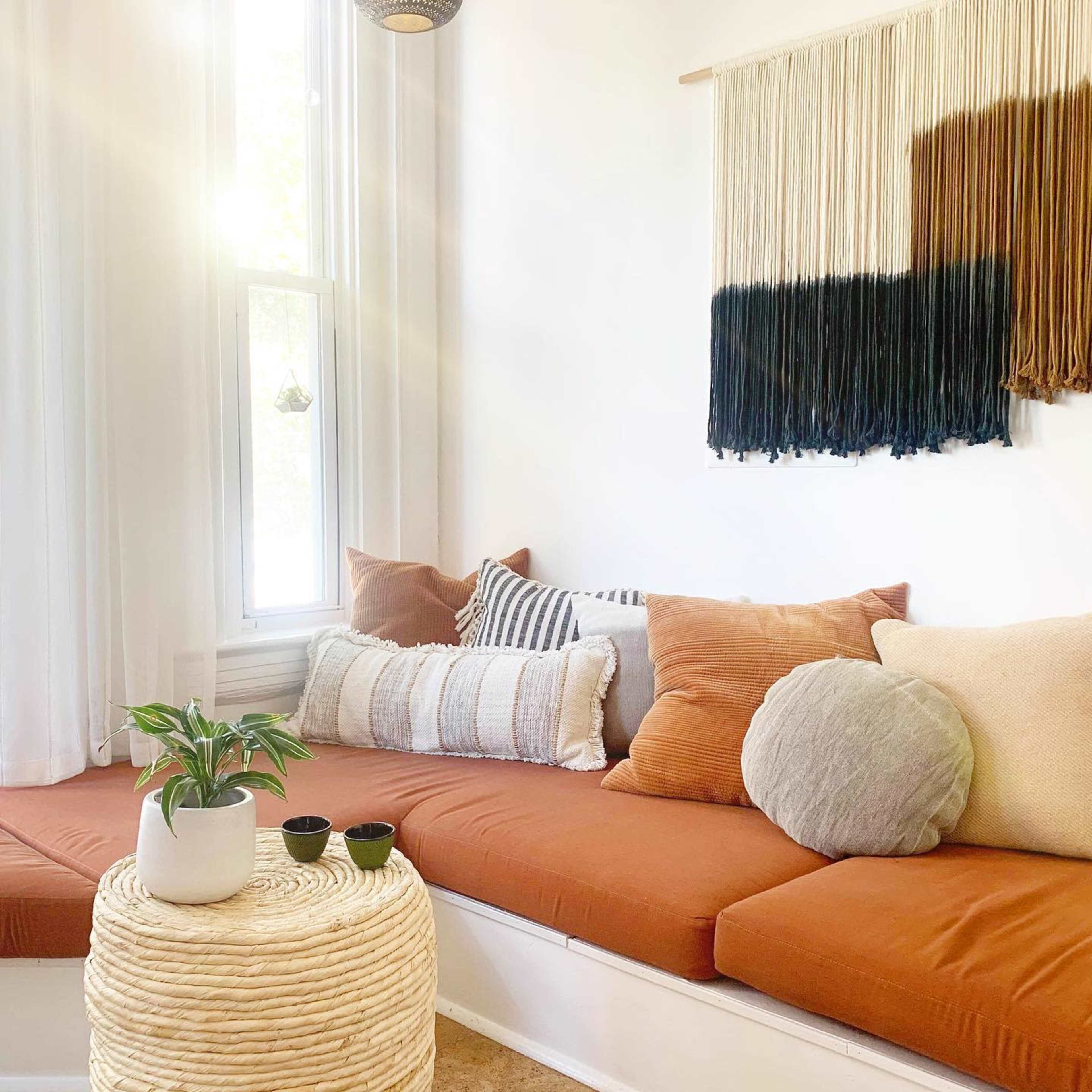 A cozy seating area features a large, rust-colored cushion with various decorative pillows and a textured wall hanging above, accompanied by a small round table and a houseplant.