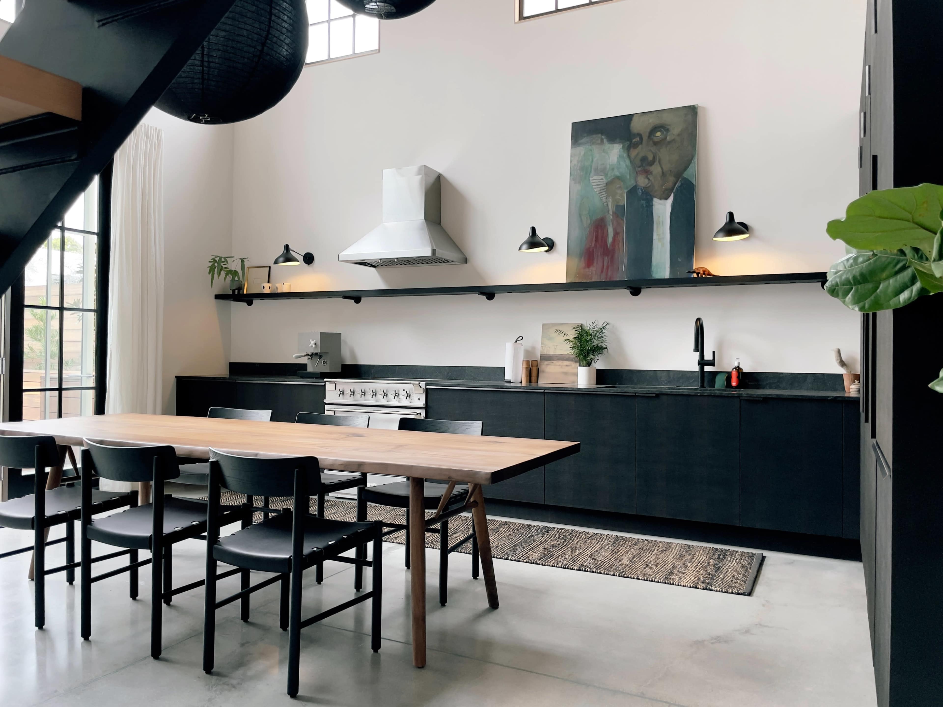 A modern kitchen features a long wooden dining table, black cabinetry, and a large artwork on the wall, with a sleek stove and minimalist decor.