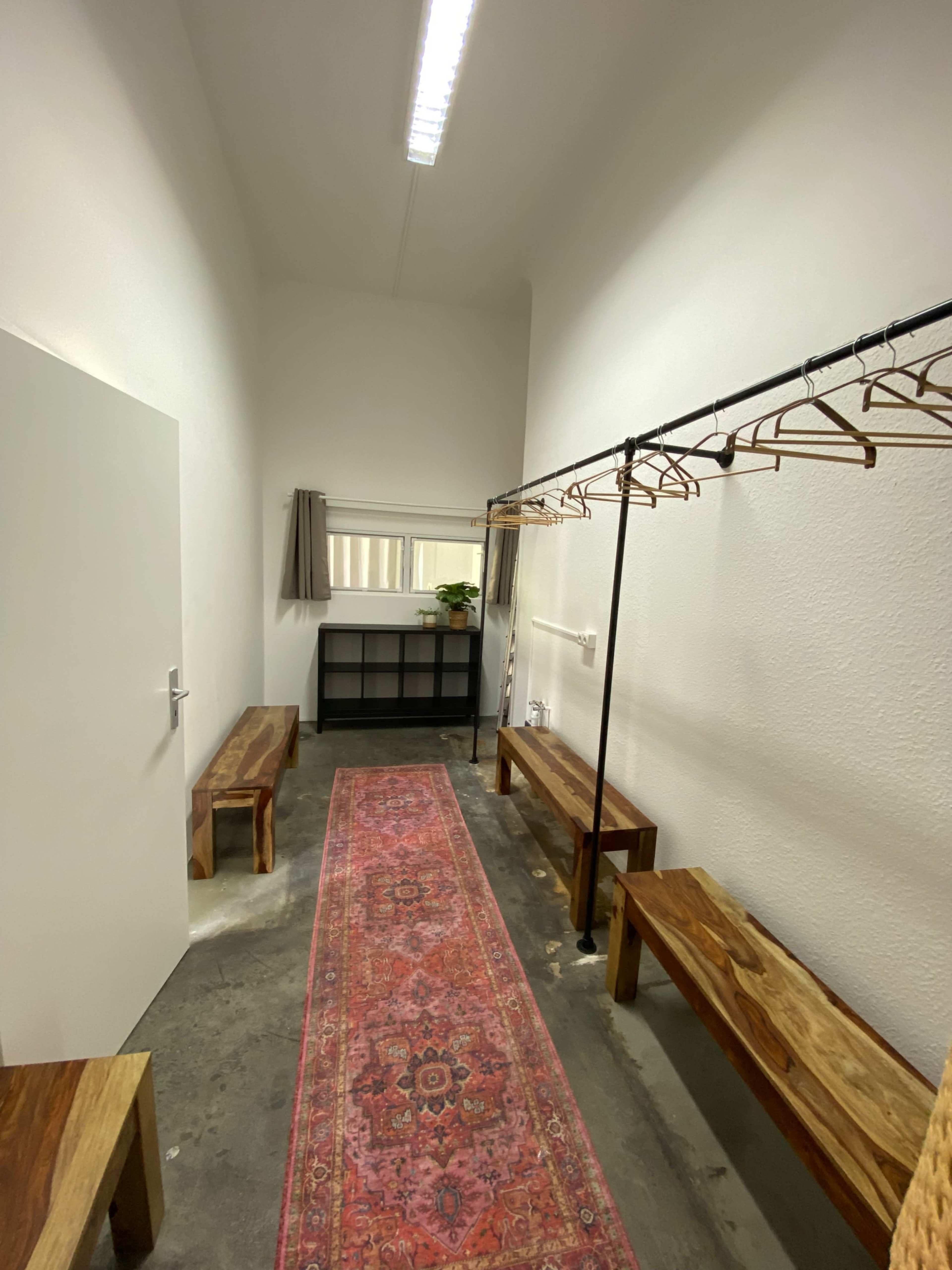 A narrow, well-lit room features wooden benches, a clothing rack with hangers, a shelf, and a decorative rug running down the center.