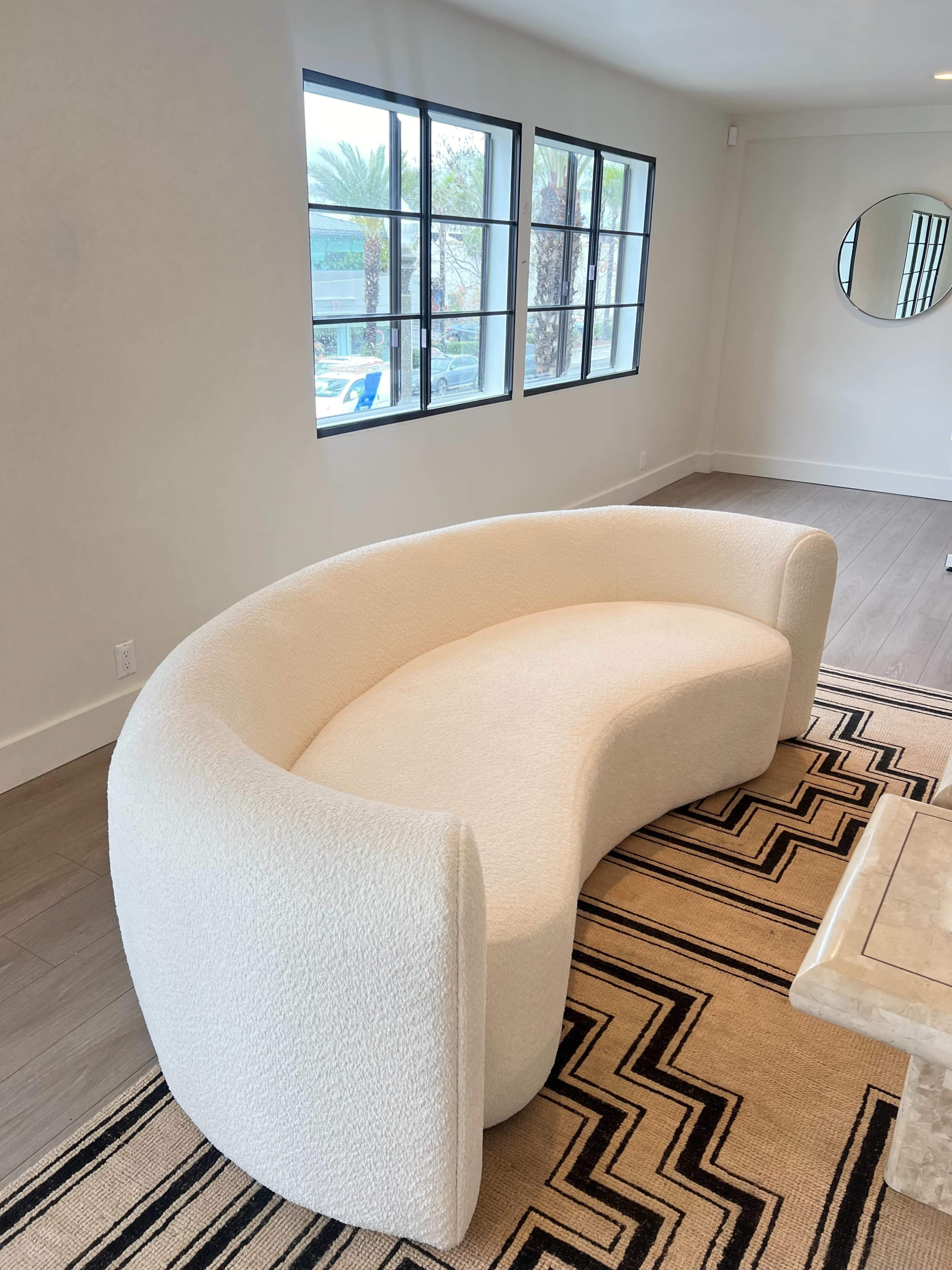 A curved, white sofa placed on a patterned rug in a well-lit room with large windows.