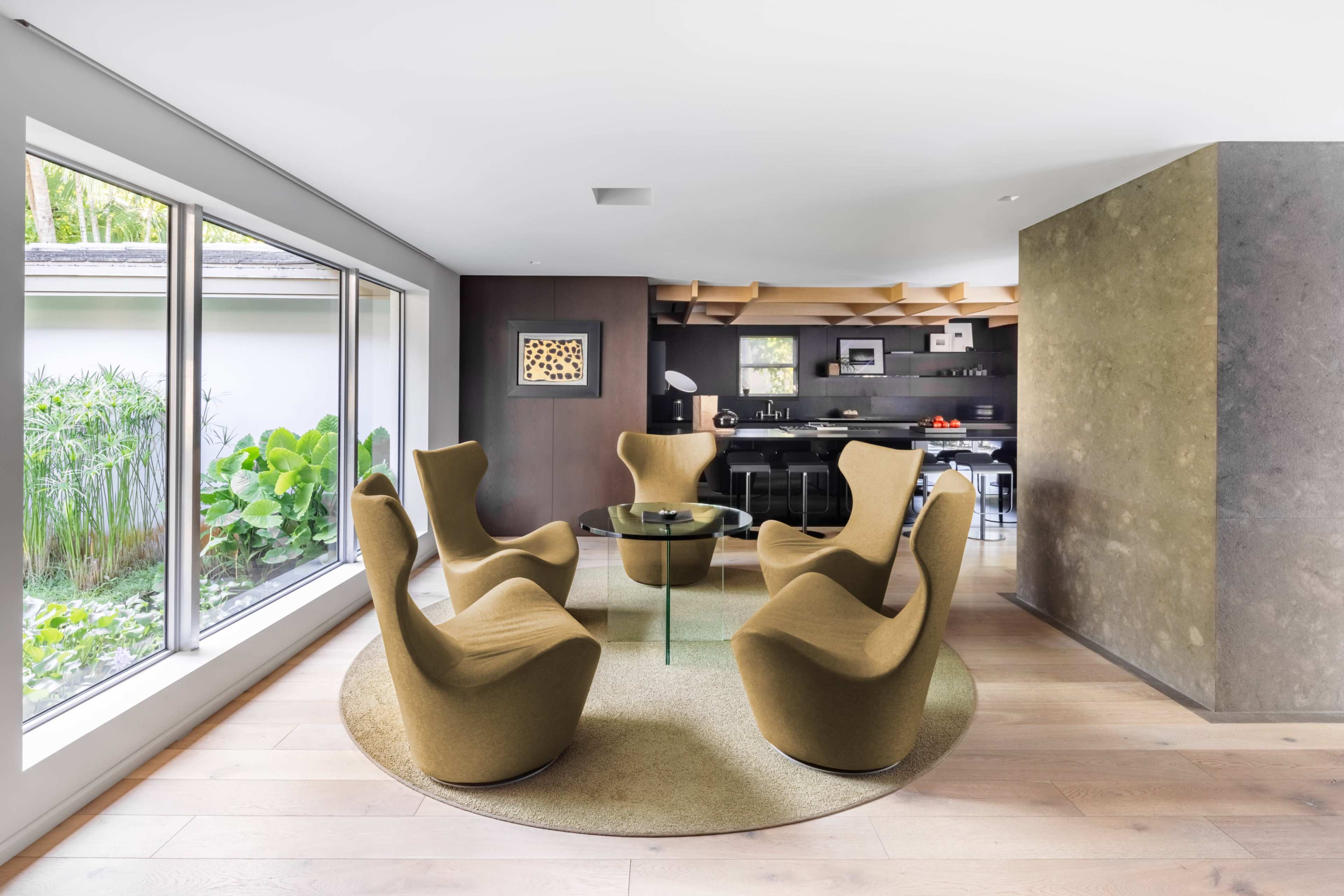 The image shows a modern living space with four brown chairs arranged around a glass table, flanked by large windows with views of greenery outside.
