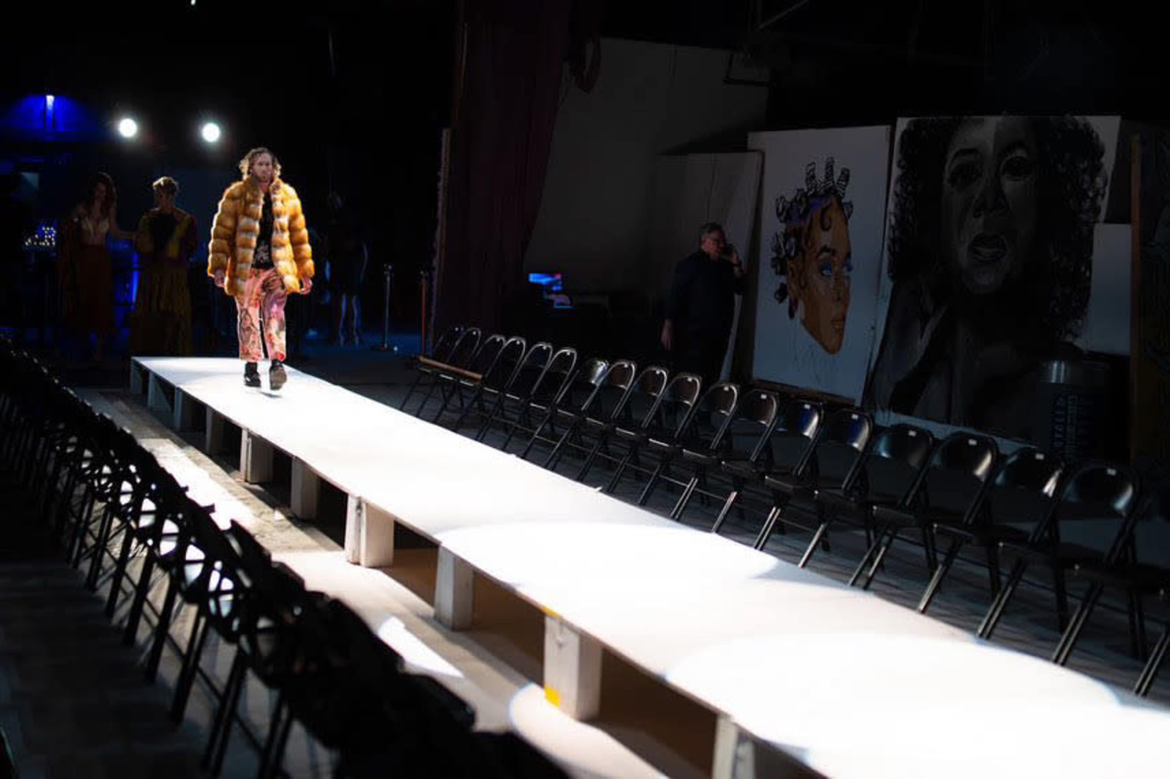 A model walks down a long, white runway lined with empty black chairs, flanked by large wall murals in a dimly lit space.