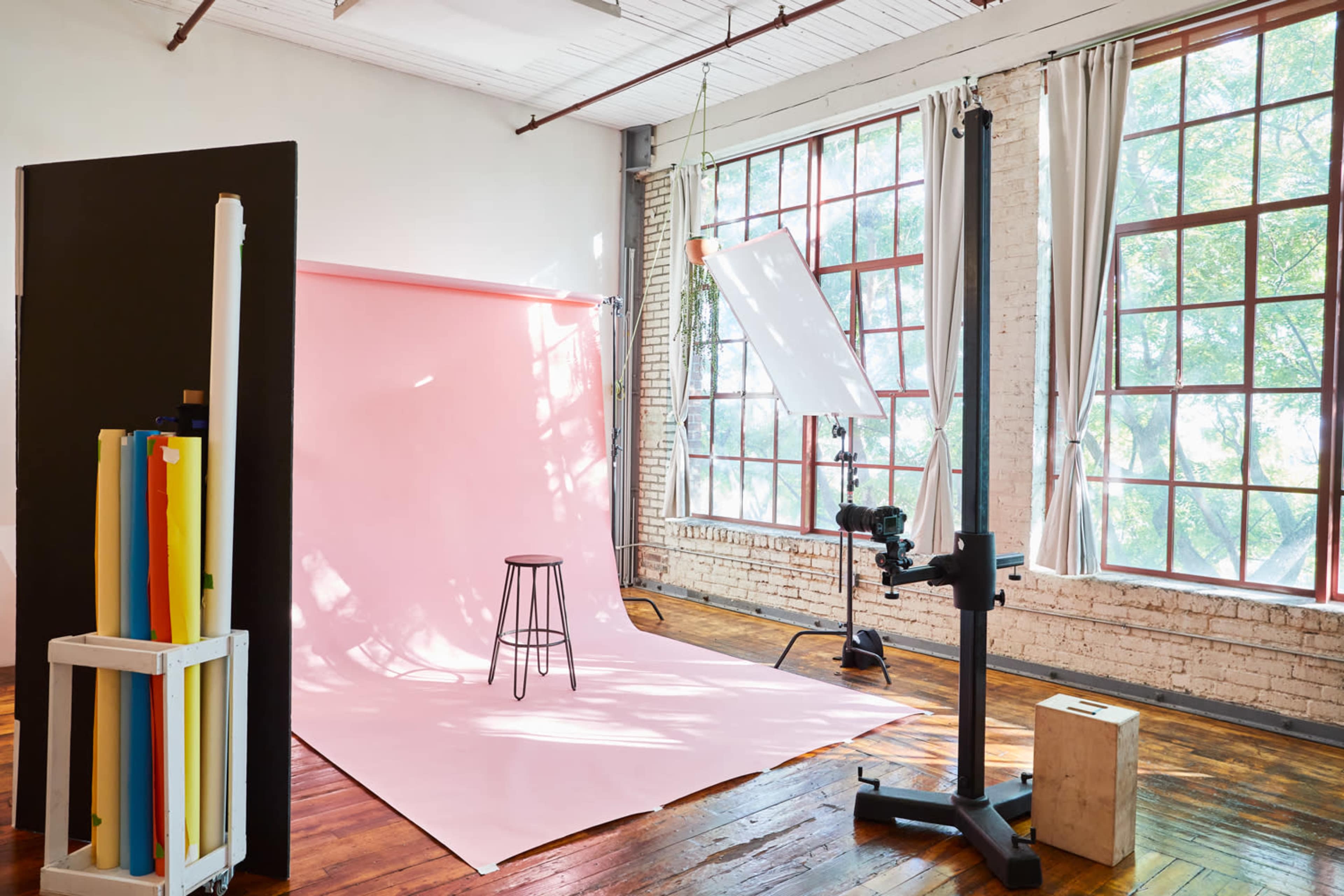 A photography studio setup featuring a pink backdrop, a stool, and various lighting equipment near large windows.