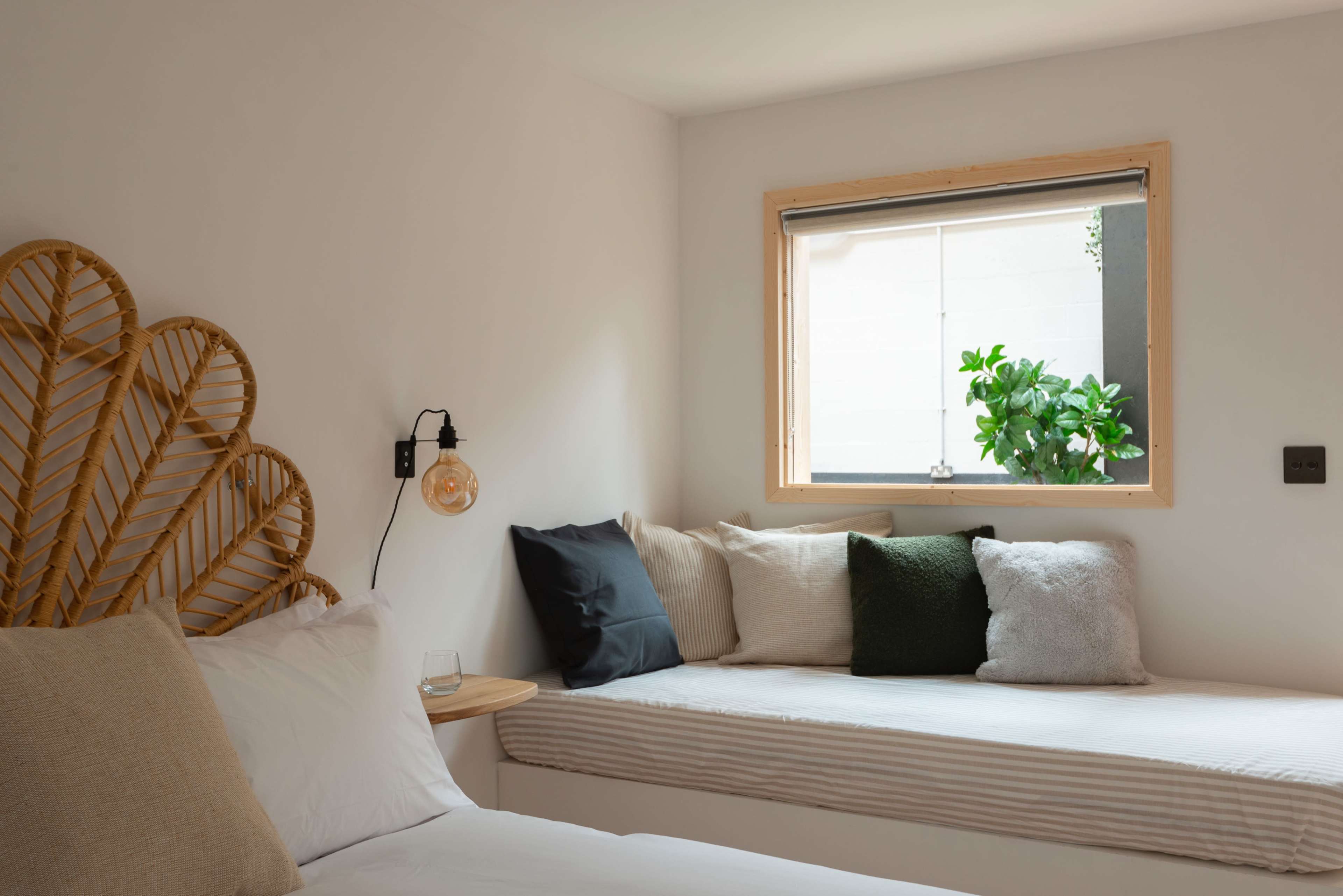 A brightly lit room features a bed with a rattan headboard and a window seat adorned with cushions.