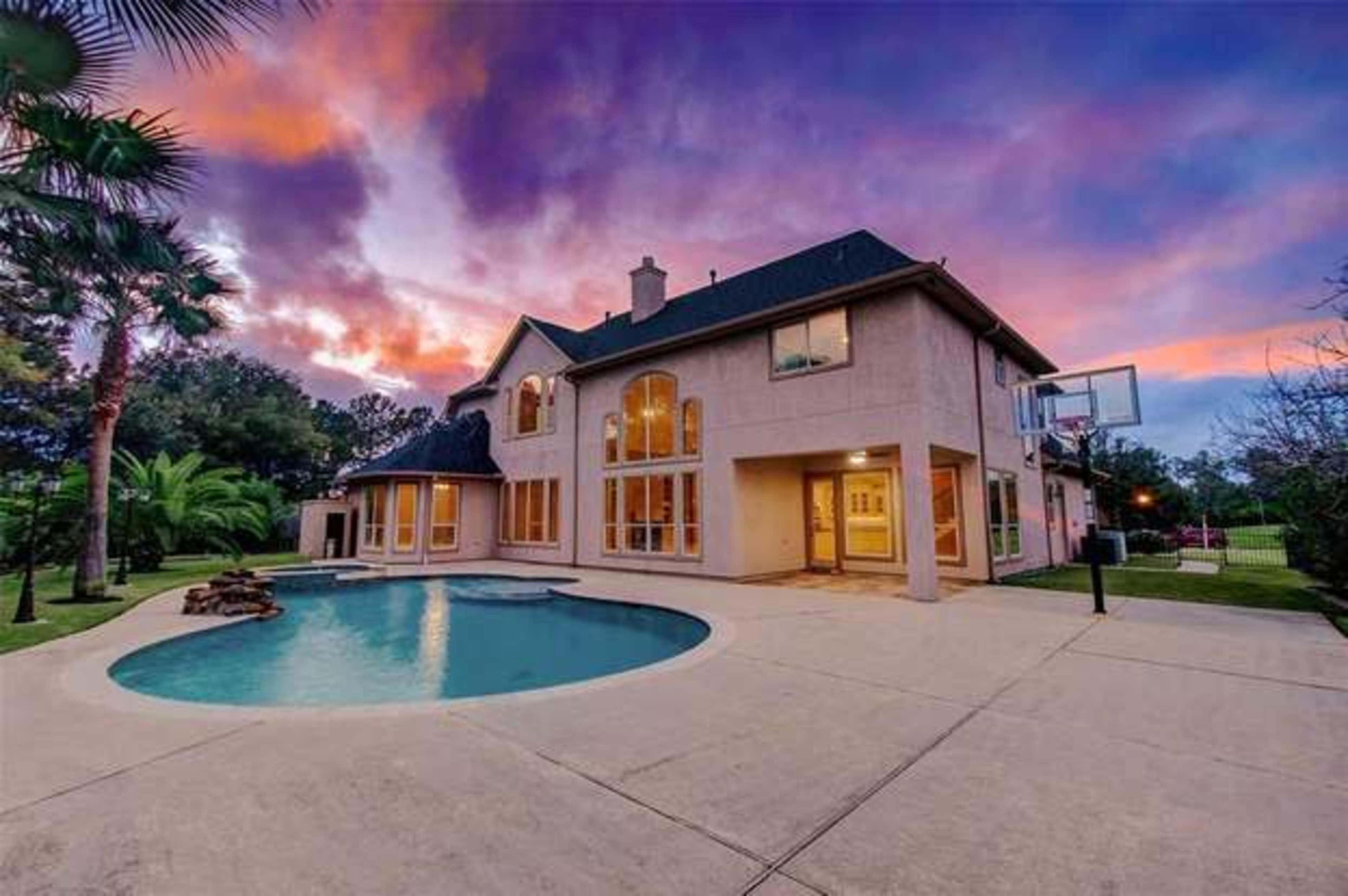 A two-story house with large windows, a swimming pool, and a basketball hoop, set against a vibrant sunset sky.
