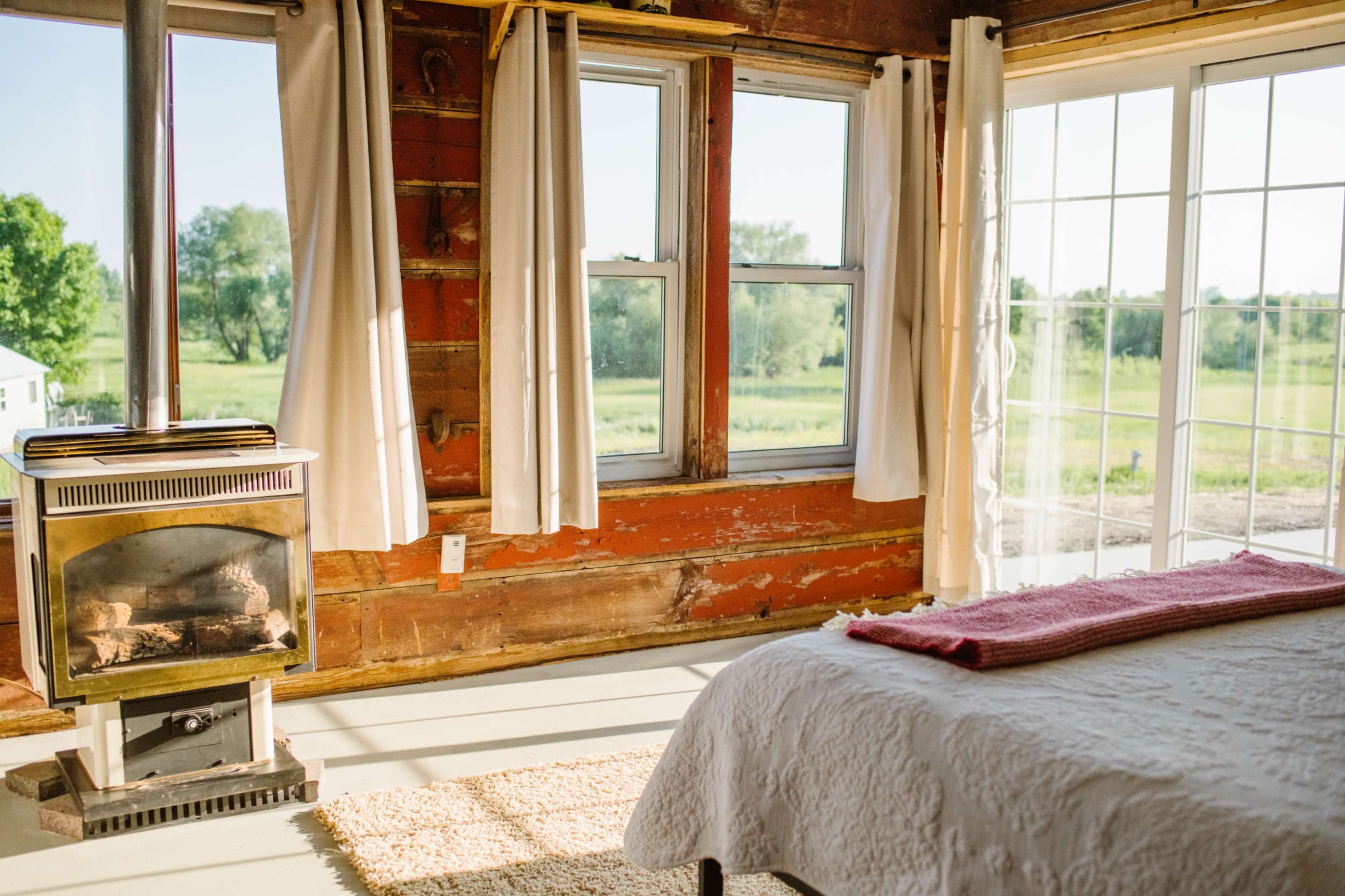 A cozy room features a bed with a pink throw, a wood stove, and large windows that open to a green landscape.