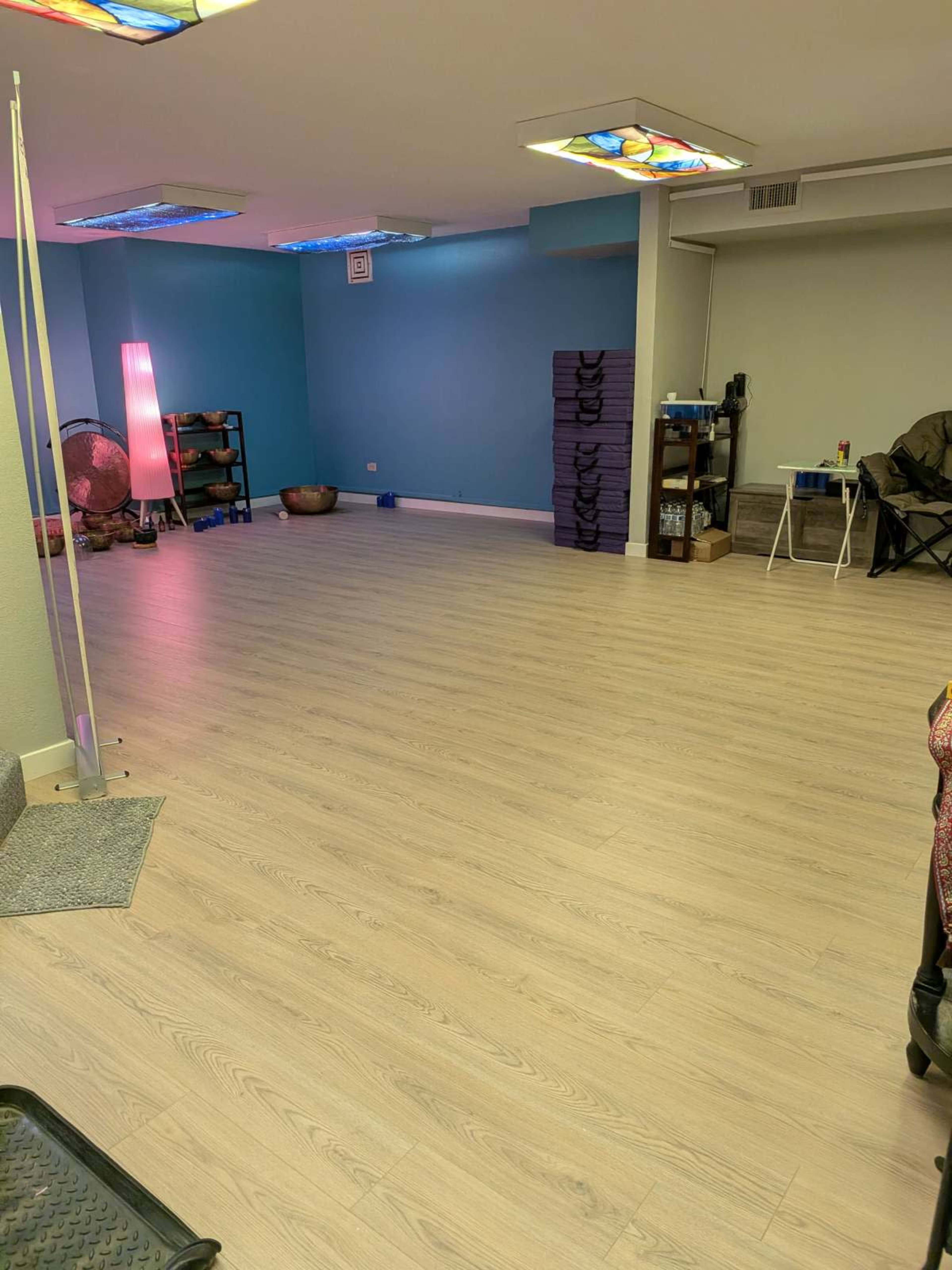 The image shows a spacious, empty room with light-colored flooring, blue walls, and a few pieces of furniture, including storage shelves and a folding chair.