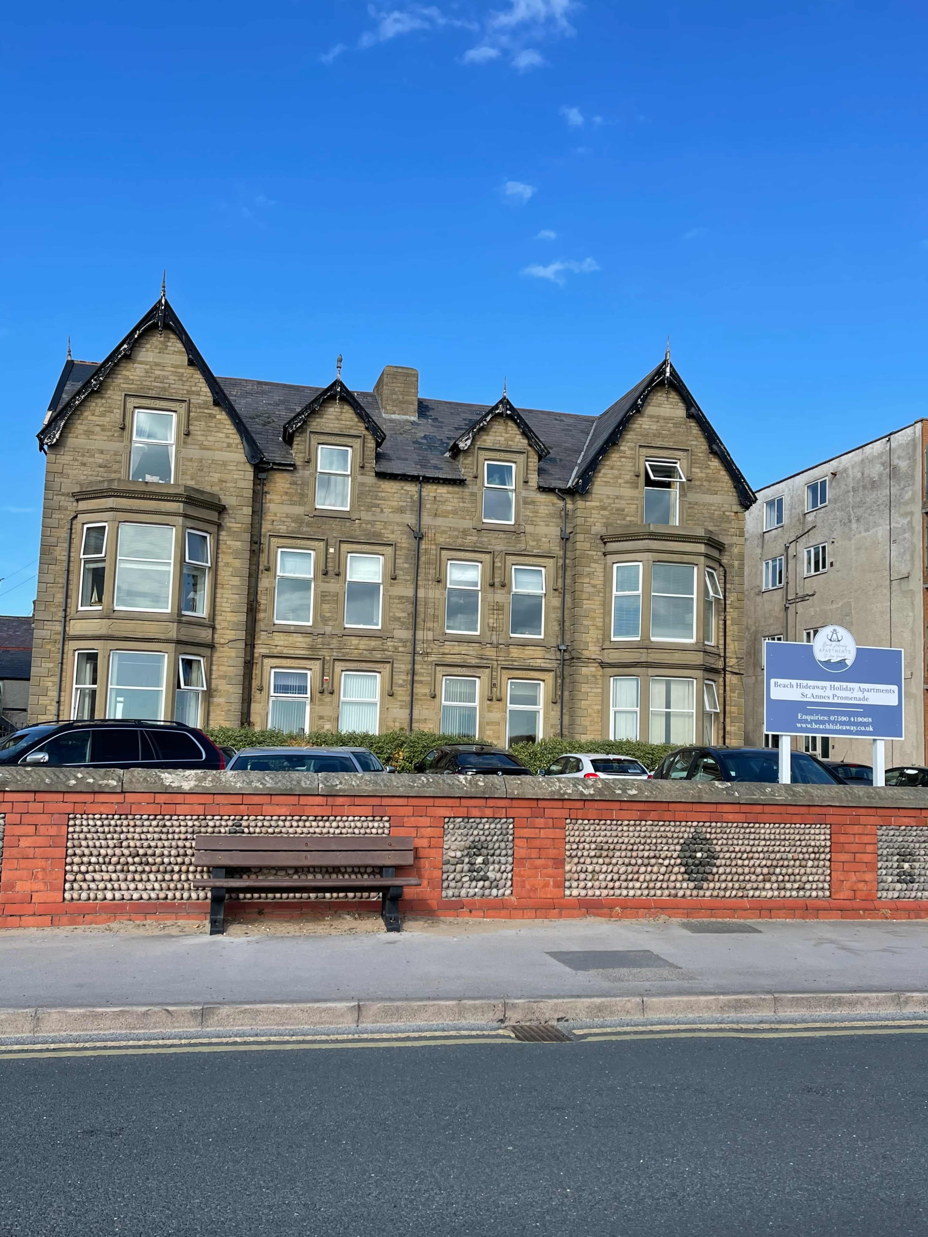 A large stone building with multiple windows and peaked roofs stands beside a sign and a brick wall.