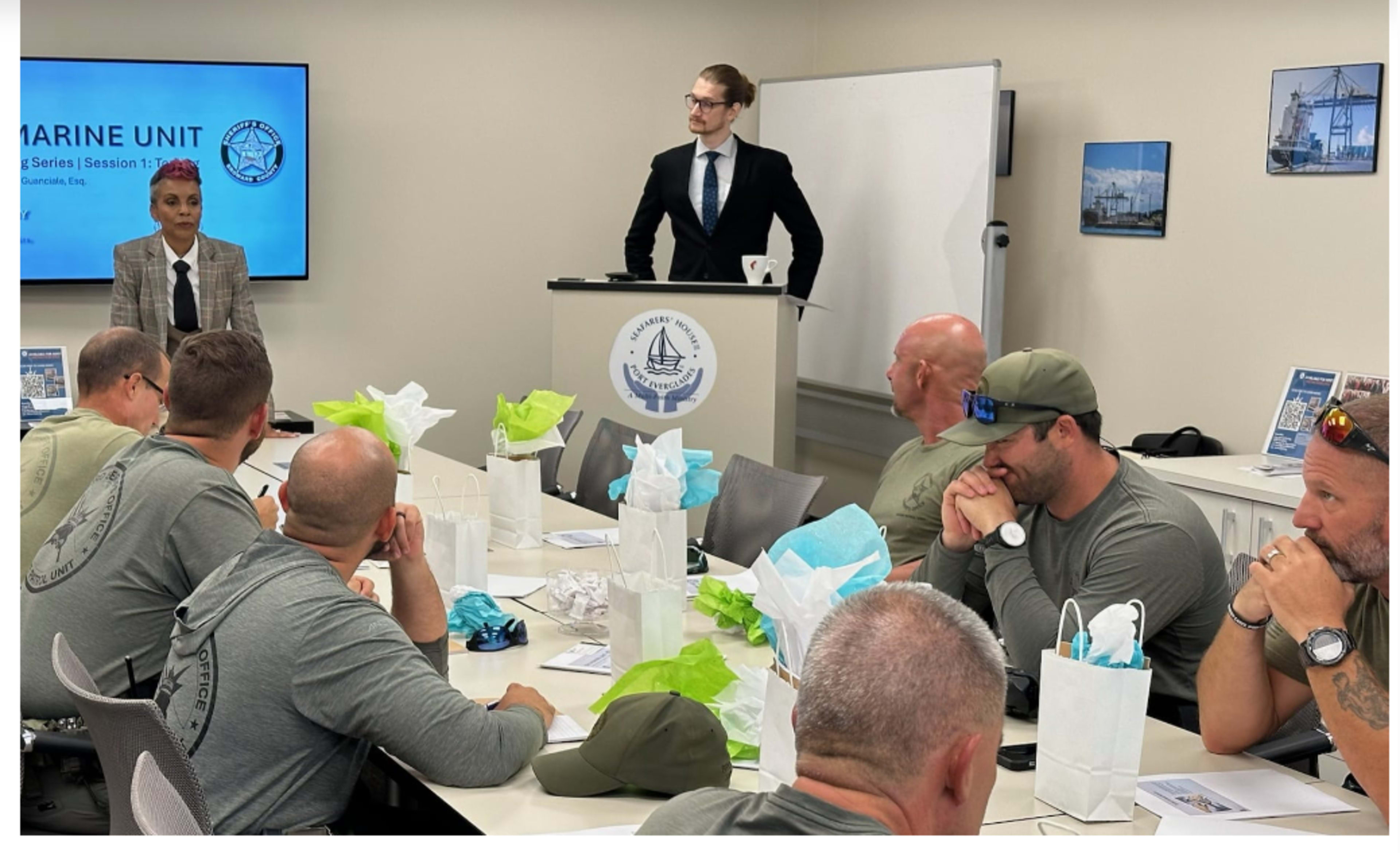 A professional meeting is taking place in a conference room where a presenter stands beside a seated audience, with attendees in matching gray shirts and gift bags on the tables.