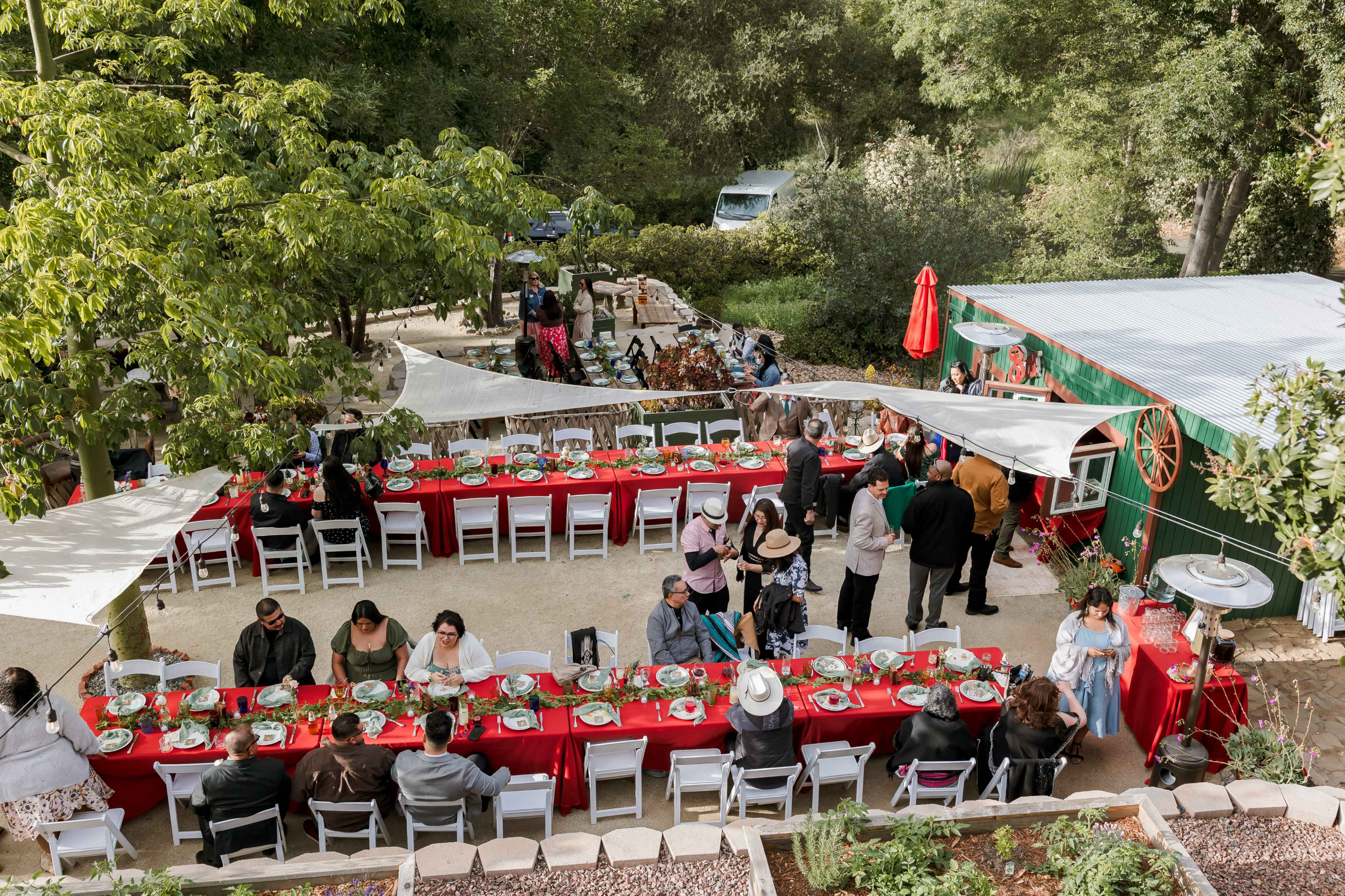 A large outdoor gathering features multiple tables adorned with red tablecloths, surrounded by guests enjoying a meal in a garden setting.