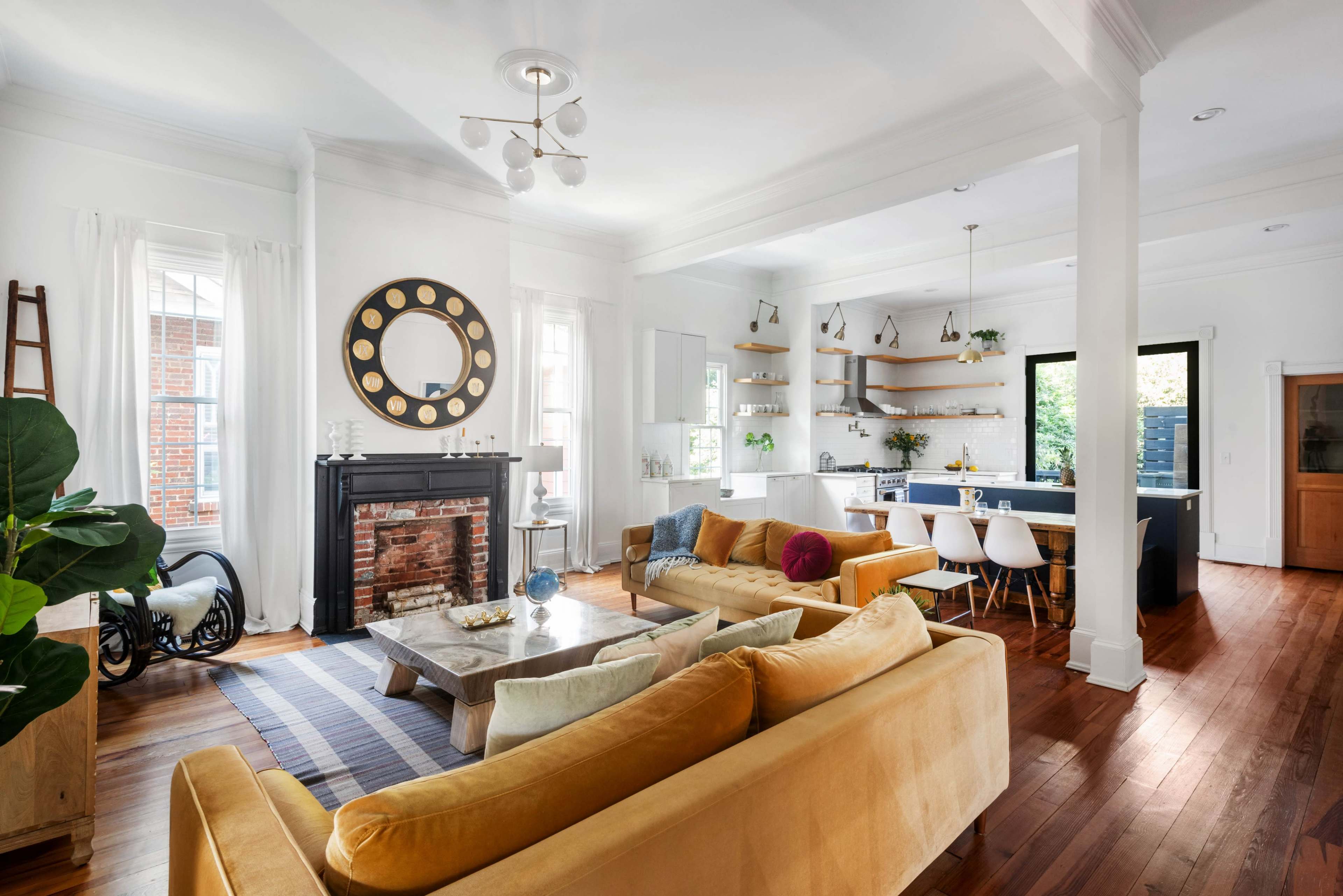 The open living space features a comfortable seating area with yellow sofas, a fireplace with a large round mirror above it, and a modern kitchen visible in the background.