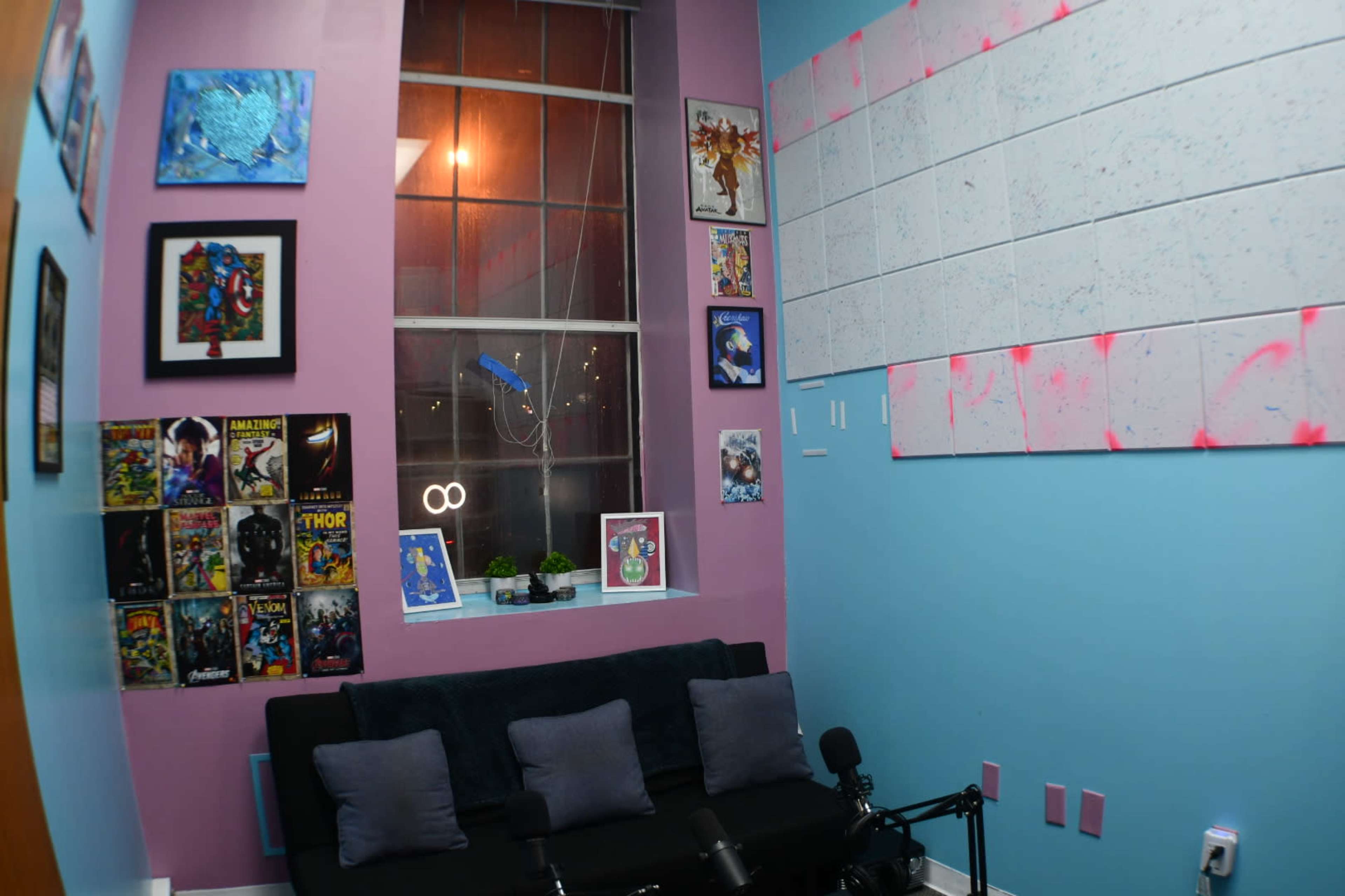 A cozy room featuring a black sofa with gray cushions, walls painted in pink and blue, and various framed artworks and comic book covers arranged on the walls.